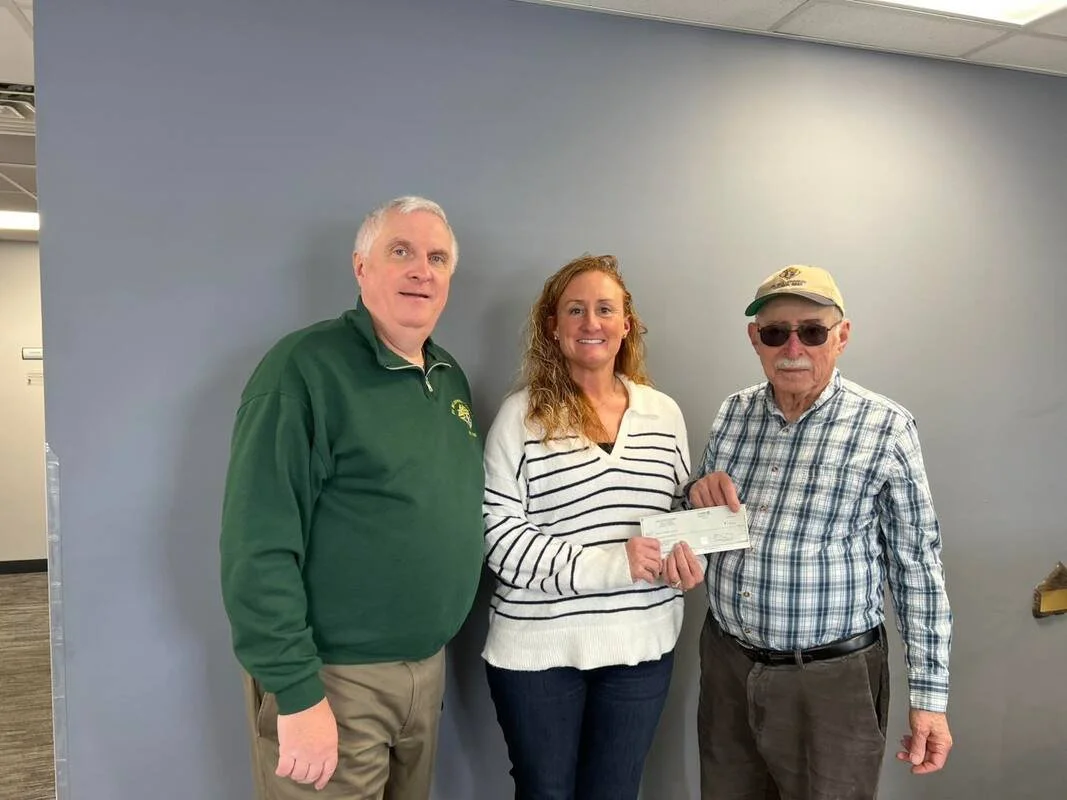 Three people standing indoors in front of a plain gray wall, holding a check. The person in the middle, a woman with curly red hair, is smiling and holding the check with both hands. The man on the right is wearing sunglasses, a beige cap, and a checkered shirt. The man on the left is wearing a green pullover and khaki pants.