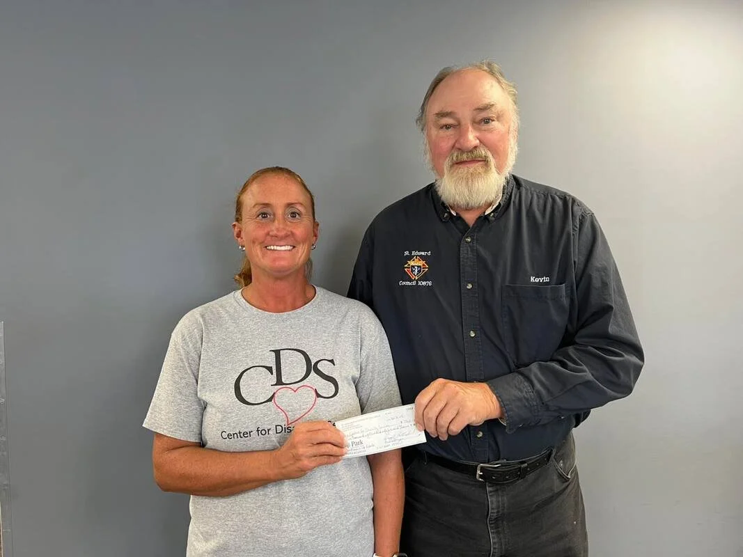A woman and a man standing together indoors, smiling at the camera. The woman is holding a check, and she is wearing a gray t-shirt with a heart design and 'Centre for Dis' printed on it. The man is wearing a black shirt with a logo and the name 'Kevin.'