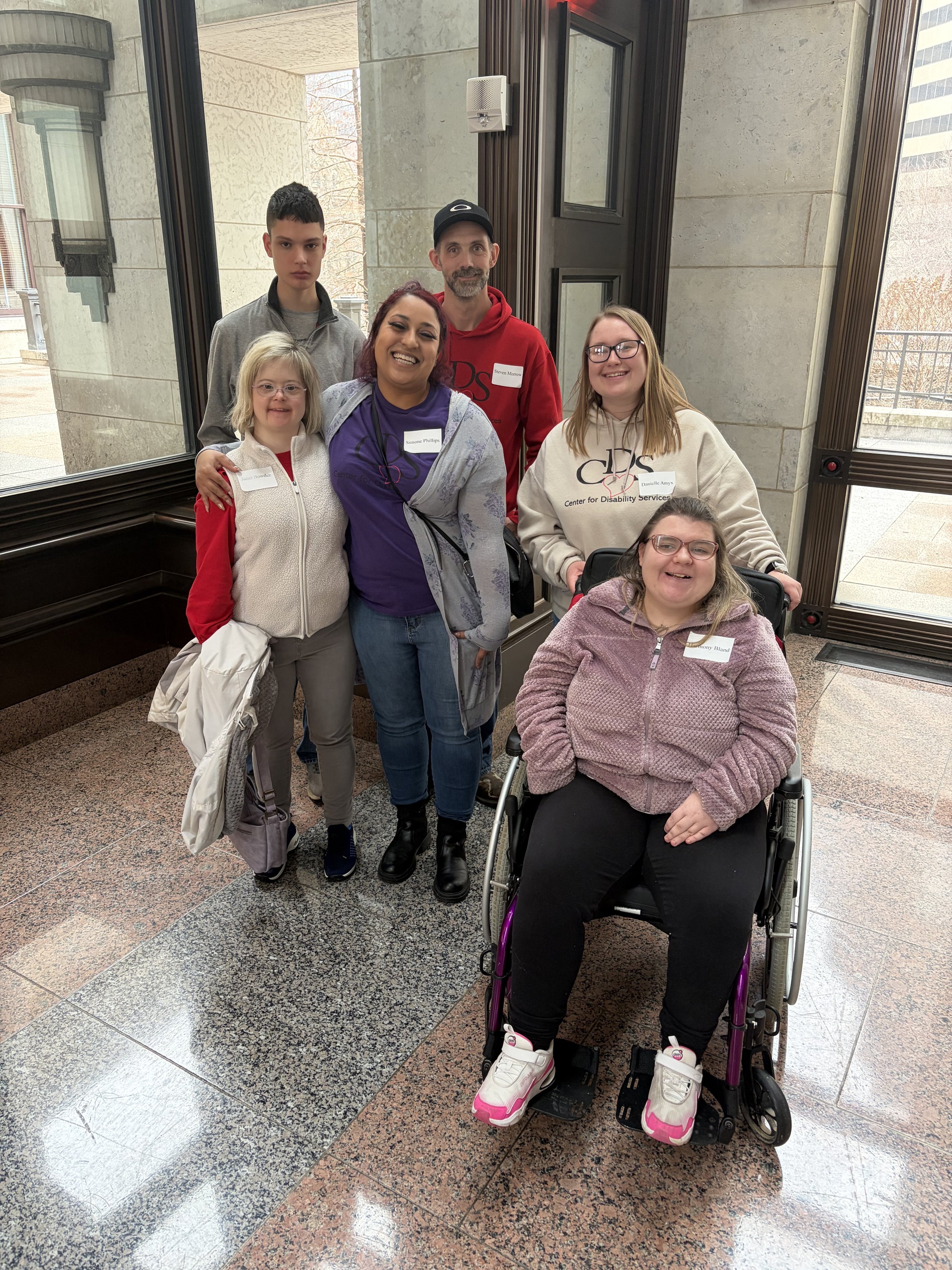 A group of six diverse people, including one in a wheelchair, smiling inside a building near a window, with exterior buildings visible outside.