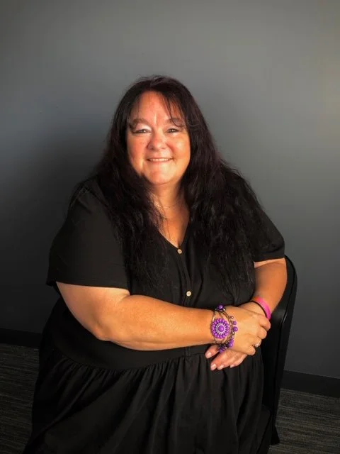 A woman with long dark hair sitting on a black chair against a gray background, smiling, wearing a black dress and purple jewelry.