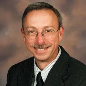 Portrait of a man with glasses, a mustache, wearing a dark suit and tie, smiling against a brown background.