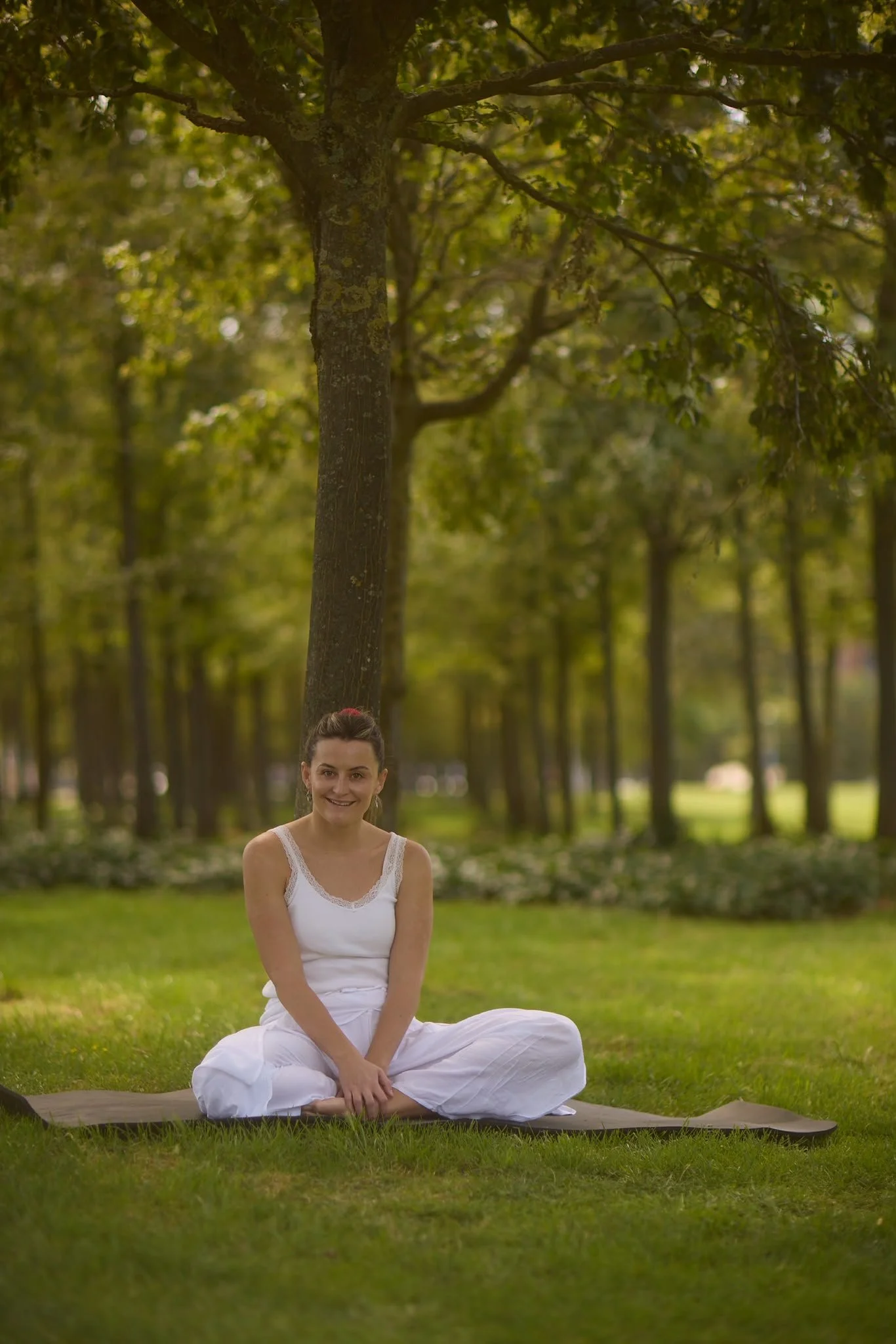 A woman sitting cross-legged on a yoga mat in a park surrounded by trees, wearing white clothing and smiling at the camera.