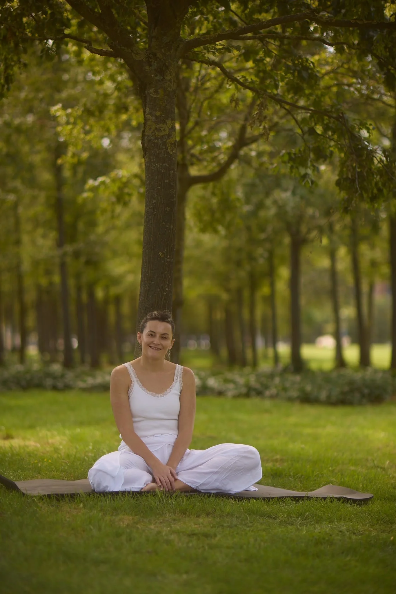 A woman in white yoga attire sits cross-legged on a yoga mat outdoors in a park with trees and grass in the background.