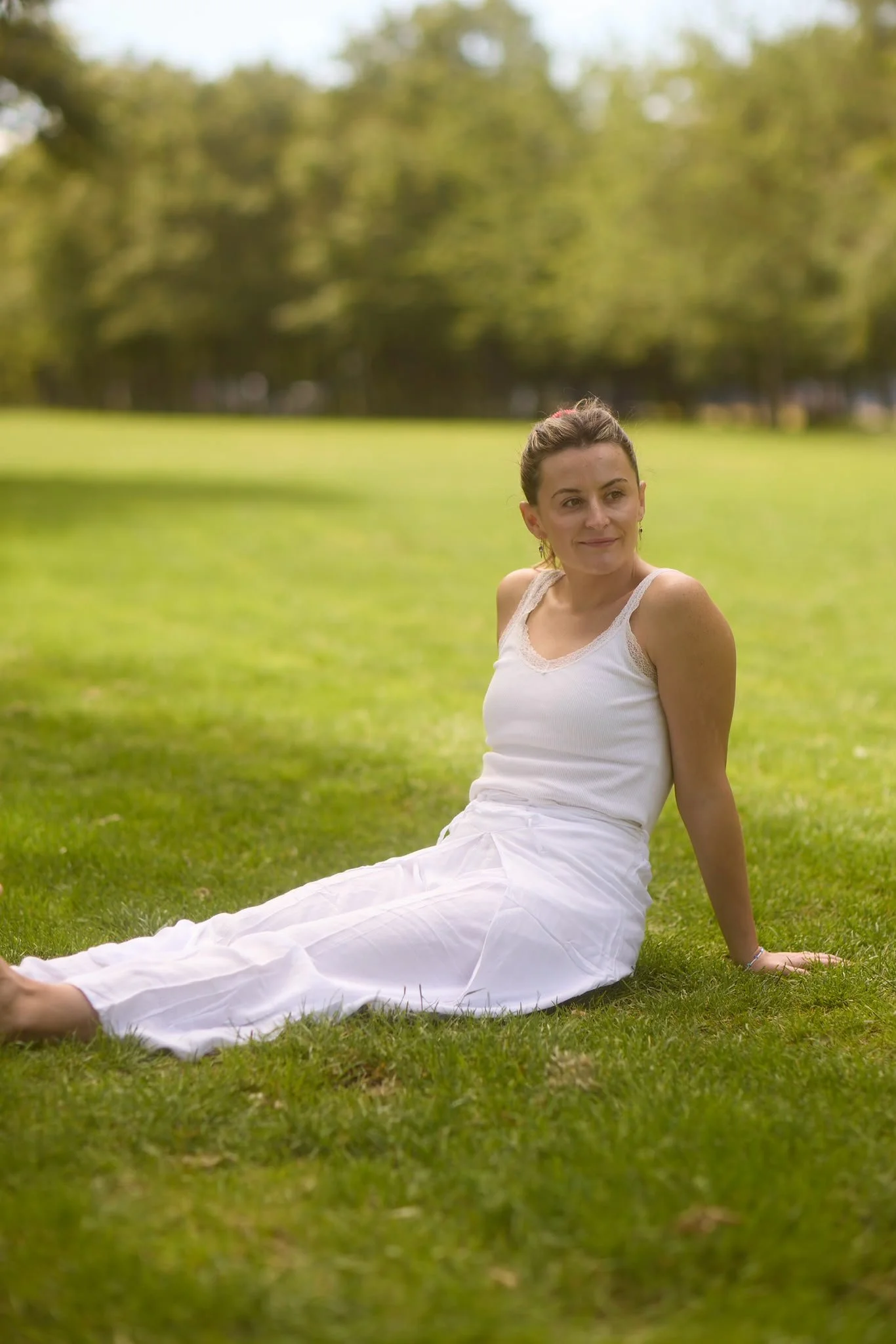 A woman in a white dress sitting on grass in a park with trees in the background.