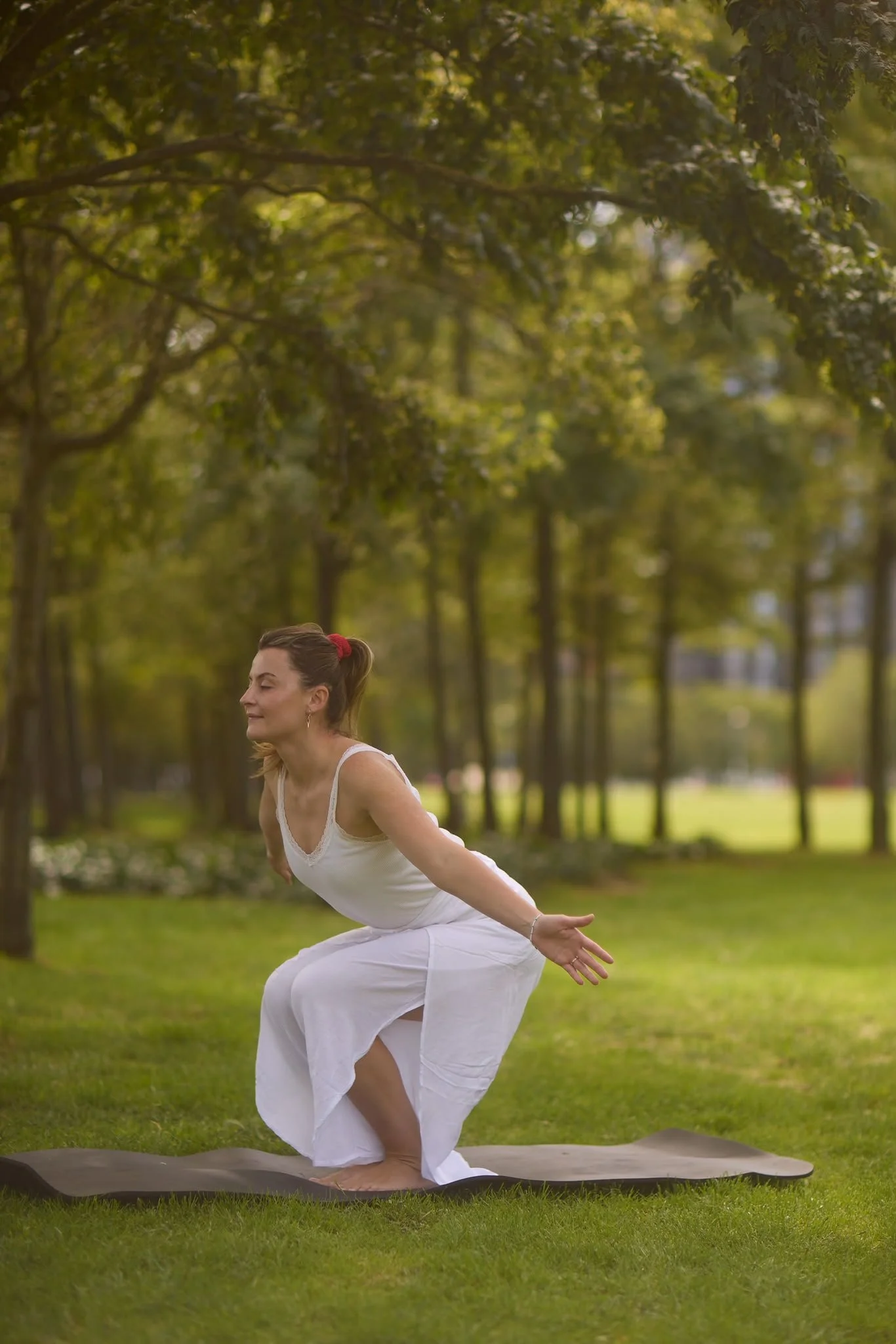 A woman in white yoga attire practicing yoga outdoors on a mat, with trees in the background.