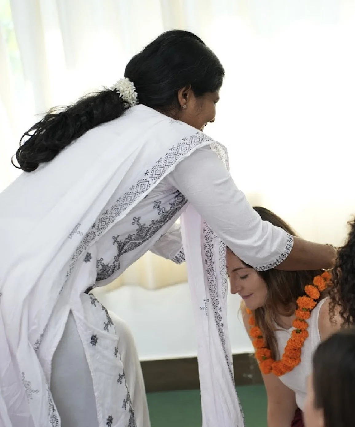 A woman in a white traditional dress with black embroidery places a flower garland on another woman who is bowing her head, in a ceremonial setting with a light-colored curtain in the background.