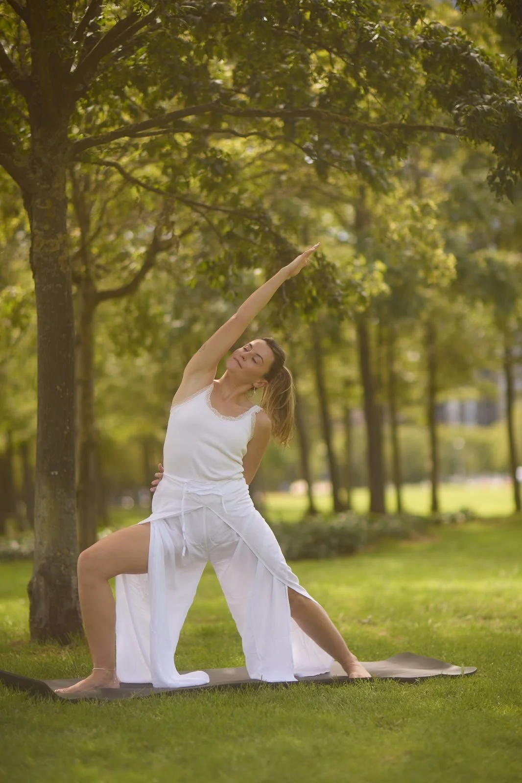 A woman practicing yoga outdoors on a yoga mat in a park surrounded by trees, wearing white clothing and performing a side stretch.