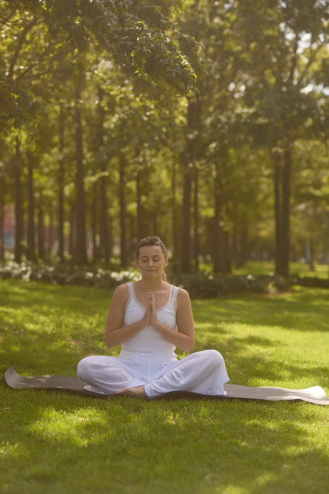 A woman practicing yoga outdoors in a park during daytime, sitting cross-legged on a yoga mat with hands in prayer position, surrounded by green grass and trees.