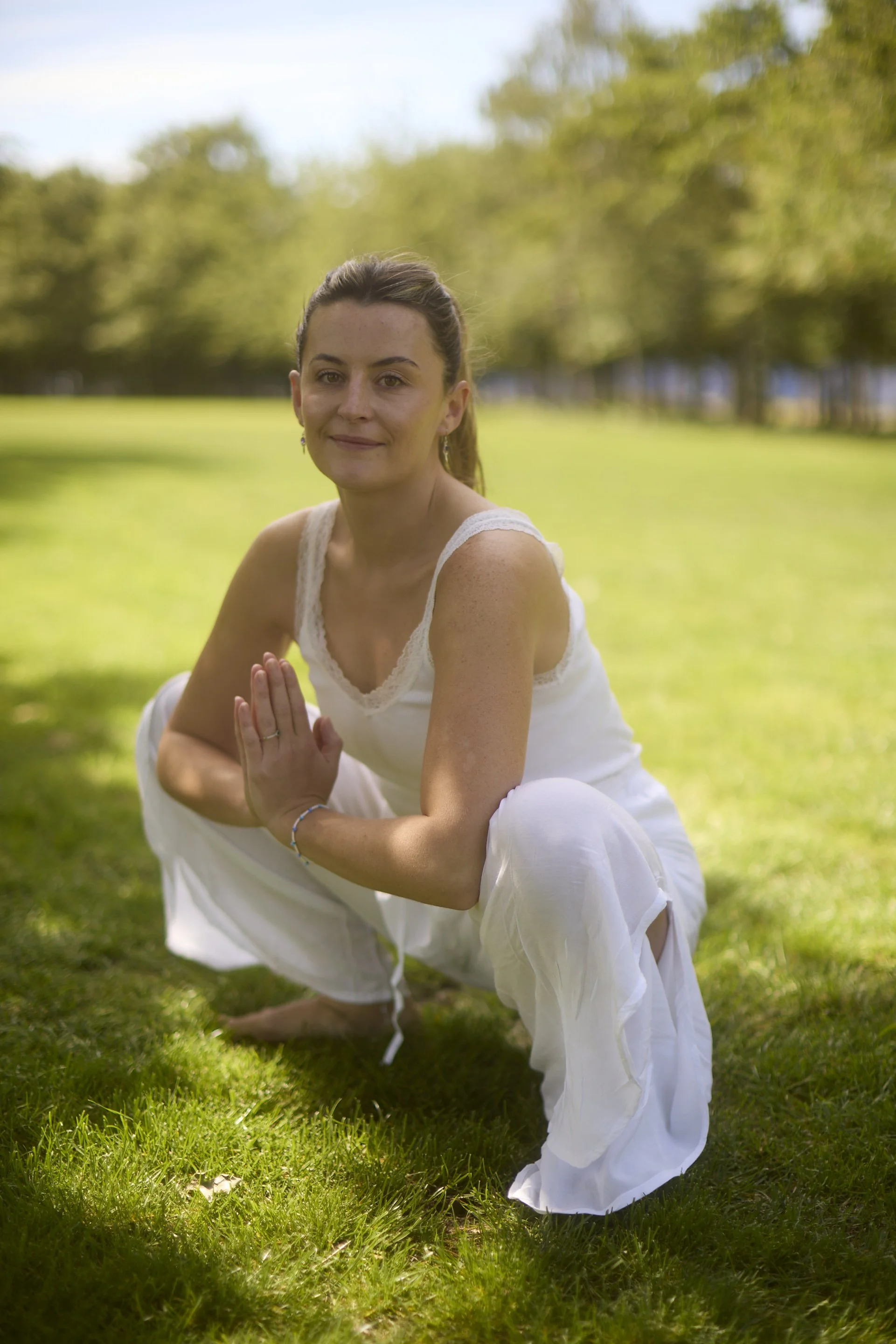 A woman practicing yoga outdoors on a sunny day, wearing a white dress, kneeling on the grass with her hands in a prayer position, surrounded by trees and a clear sky.
