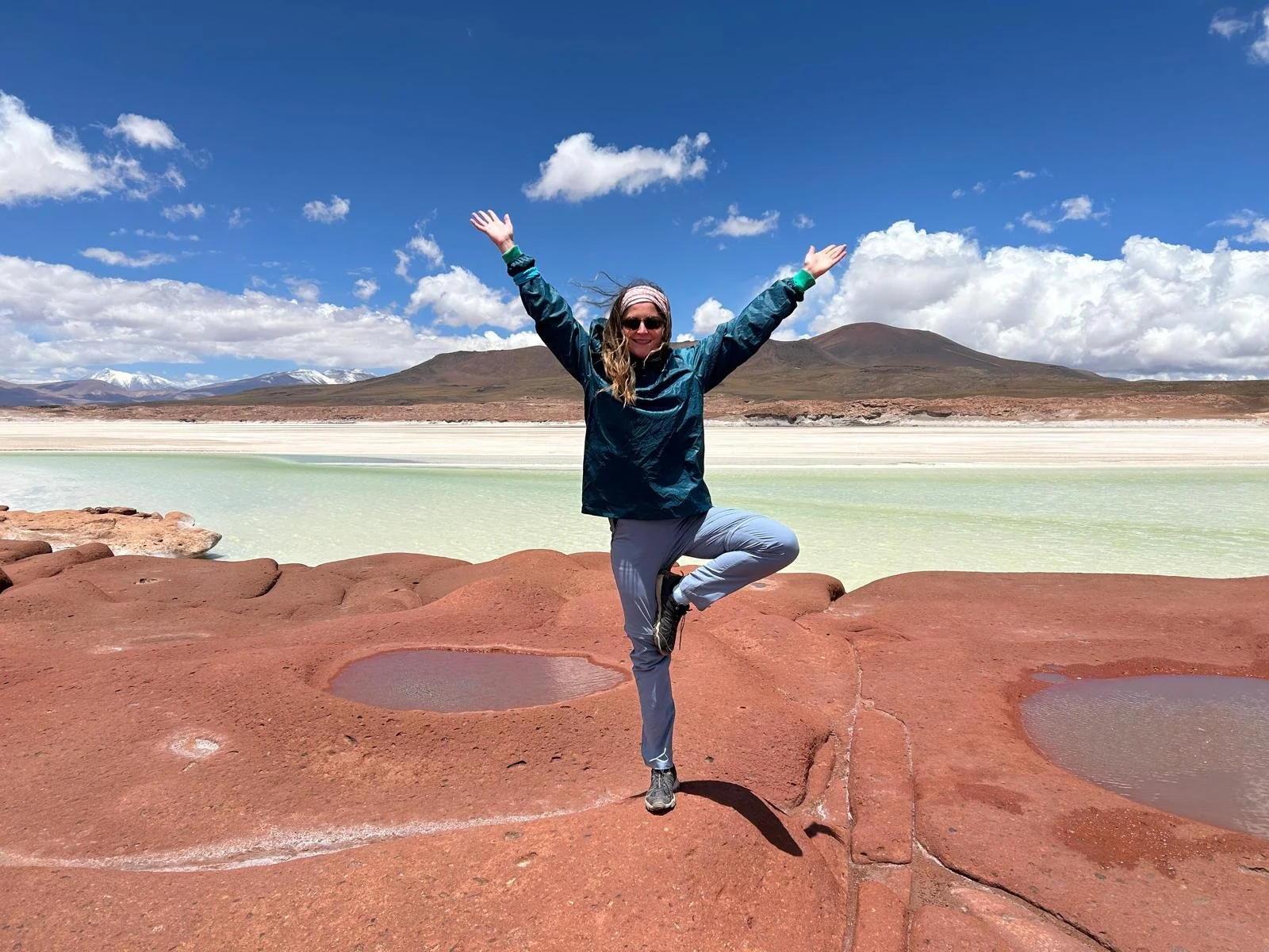 Woman in outdoor clothing performing yoga pose on rocky terrain with lakes and mountains in the background on a partly cloudy day.