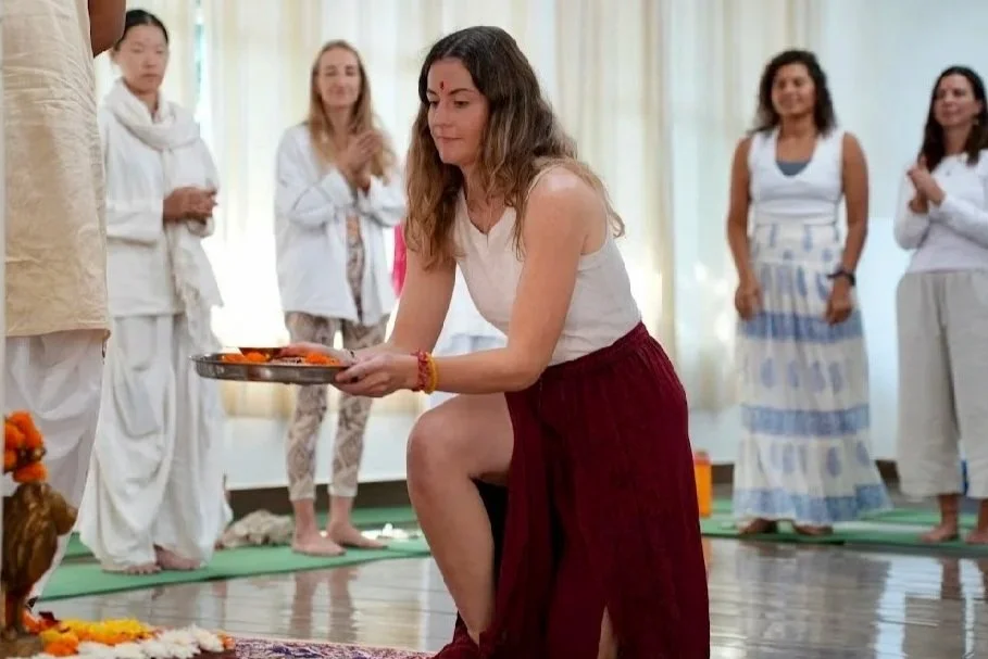 A woman in a white top and red skirt is kneeling on the floor, holding a tray of items, possibly during a cultural or religious ceremony, with several women in white and patterned clothing standing behind her in a bright room.