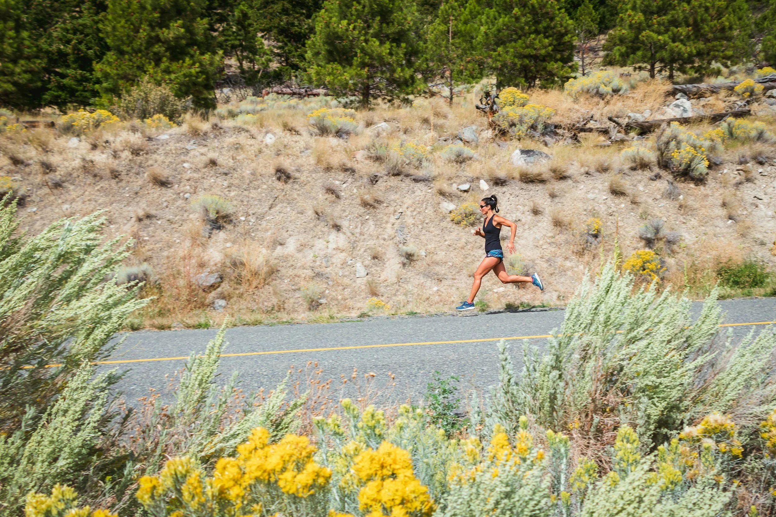 A woman running along a rural road with dry grass, desert plants, and trees in the background.