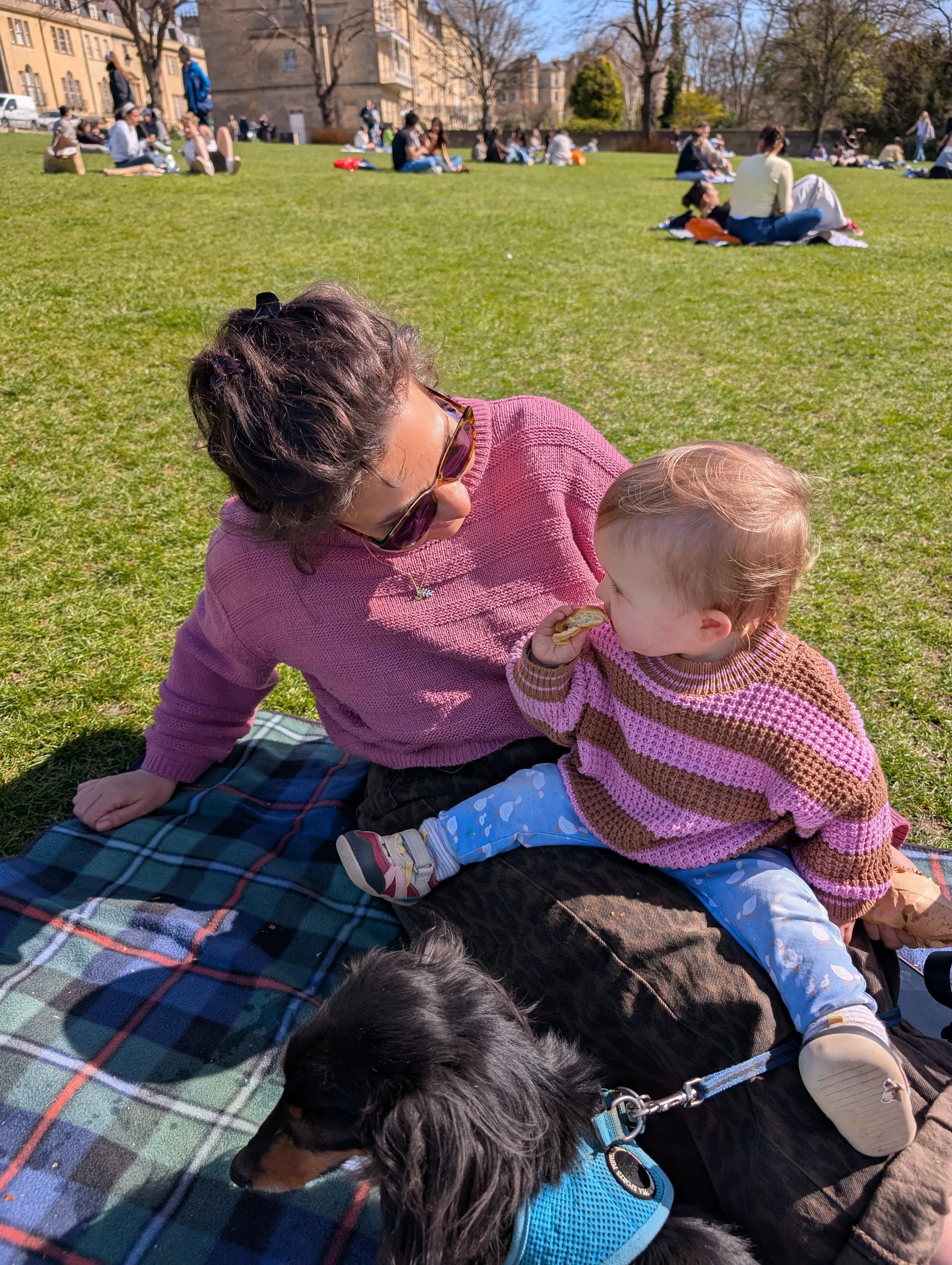 A woman in sunglasses and a pink sweater playing with a young child in a pink and brown striped sweater outdoors on a sunny day, with people sitting on a grassy area in the background.
