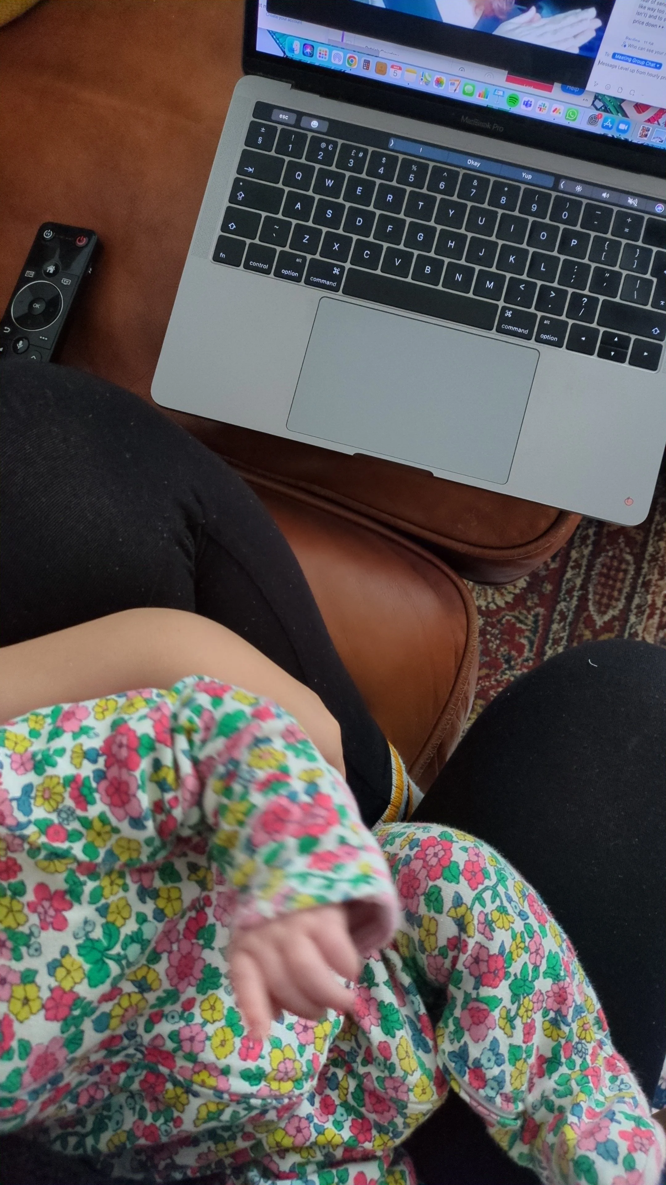 Child wearing a colorful floral dress sits on an adult's lap, with a silver laptop and TV remote on a wooden table nearby.