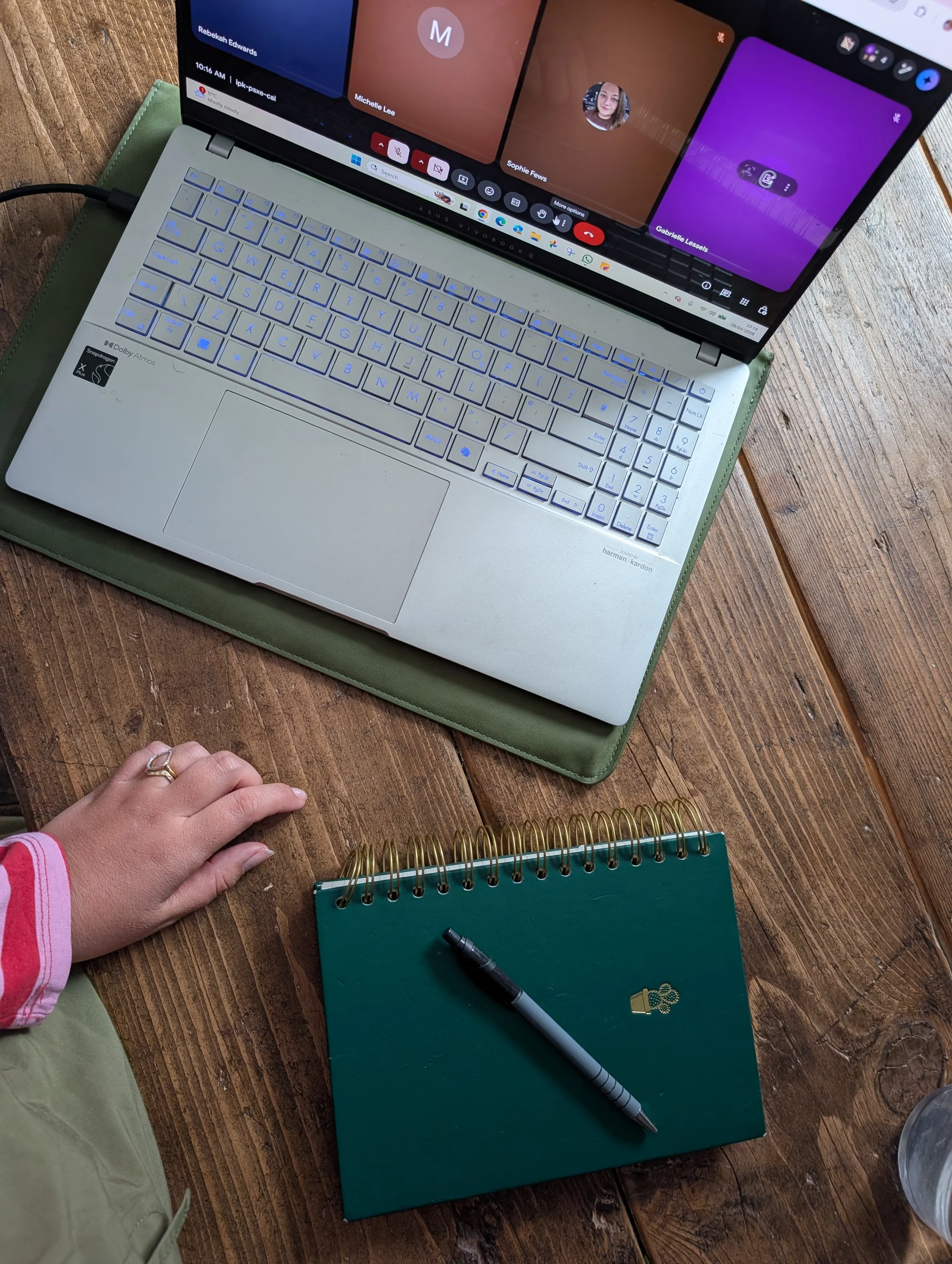 A person in a pink and white striped long sleeve shirt and khaki pants is resting their hand on a wooden desk near a green notebook with a gold emblem and a black pen. There is a silver laptop with its screen showing a video call with multiple participants, and a small green notebook is also on the desk.
