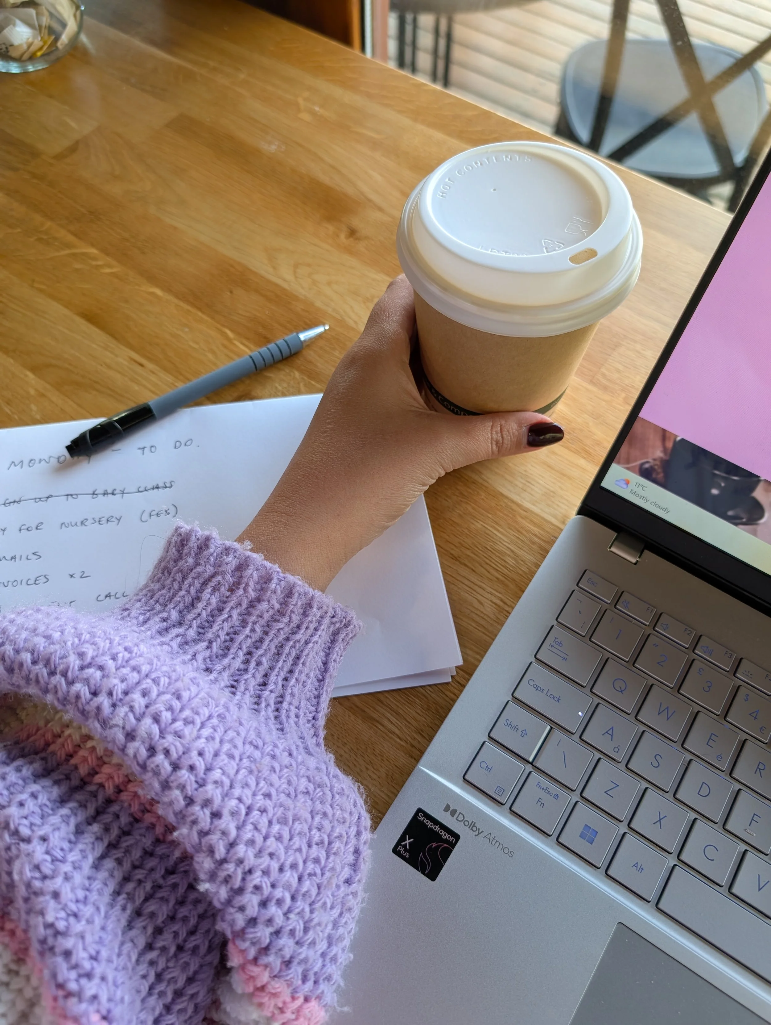 A person with painted nails and a lavender knit sweater is holding a disposable coffee cup while sitting at a wooden table. There is a notebook with handwritten notes and a pen, a laptop showing a weather forecast, and a small container with packets of sugar or sweetener on the table.