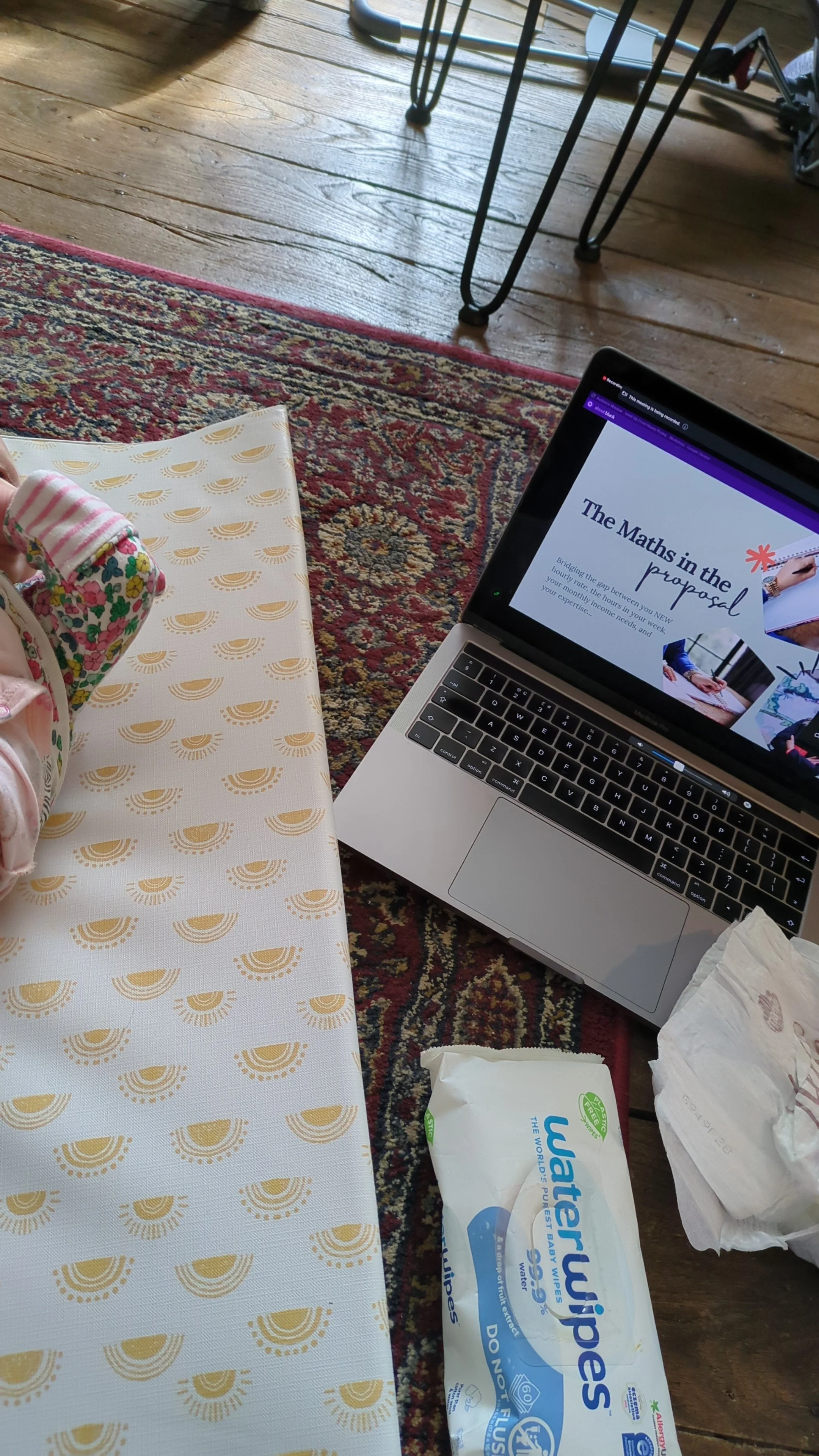 A laptop displaying a presentation titled "The Math in the Proposal" on a wooden floor next to a patterned rug, with a pack of WaterWipes baby wipes, a tissue, and someone's arm with a sleeve partially visible, while a small table with metal legs and a blurred background are also seen.