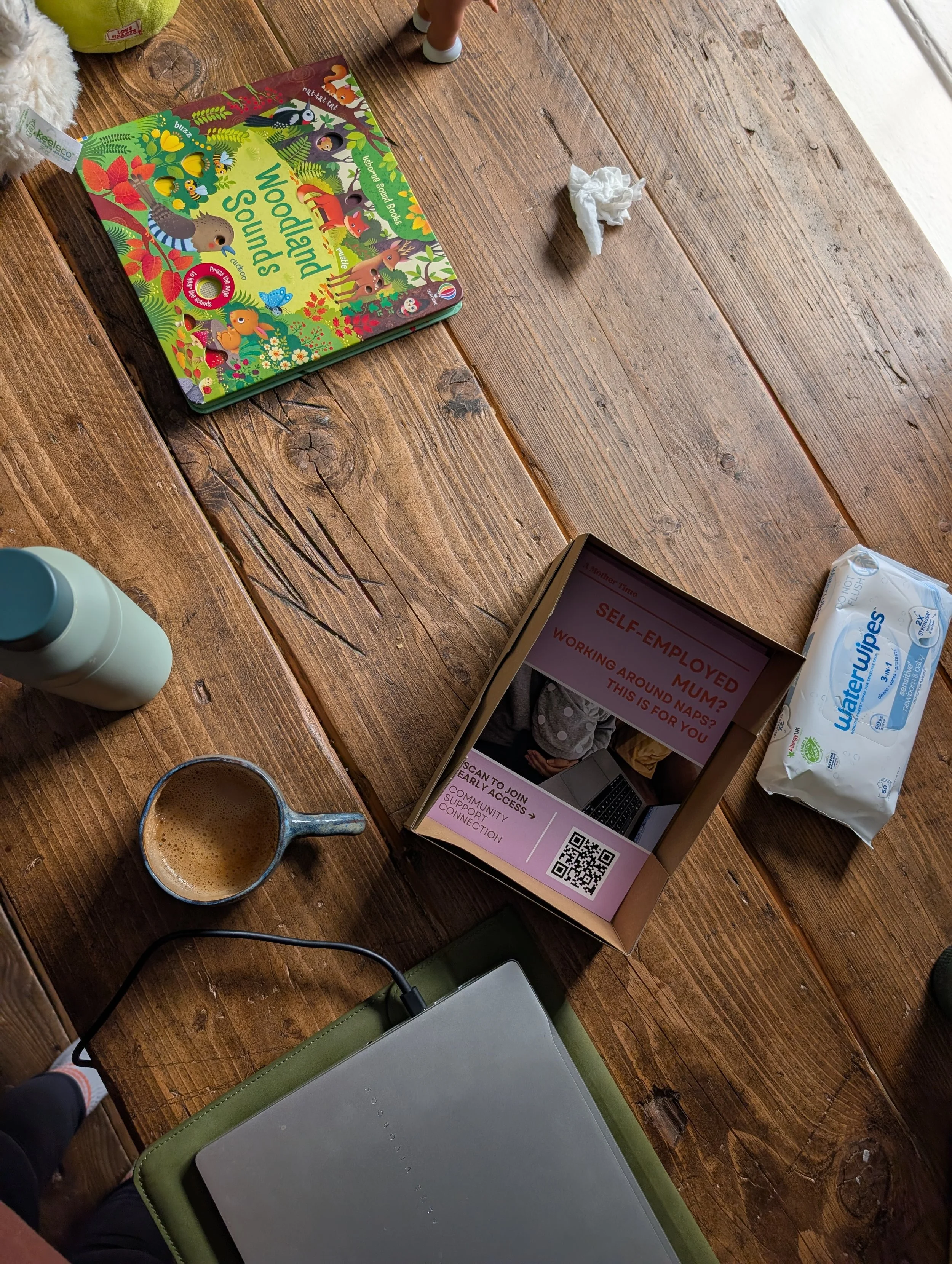A wooden table with a colorful children's book titled 'Wild Animal Sounds,' a tissue packet, a cup of coffee, a water bottle, a laptop, and a tablet showing a self-employment information page. A person's feet are partially visible.