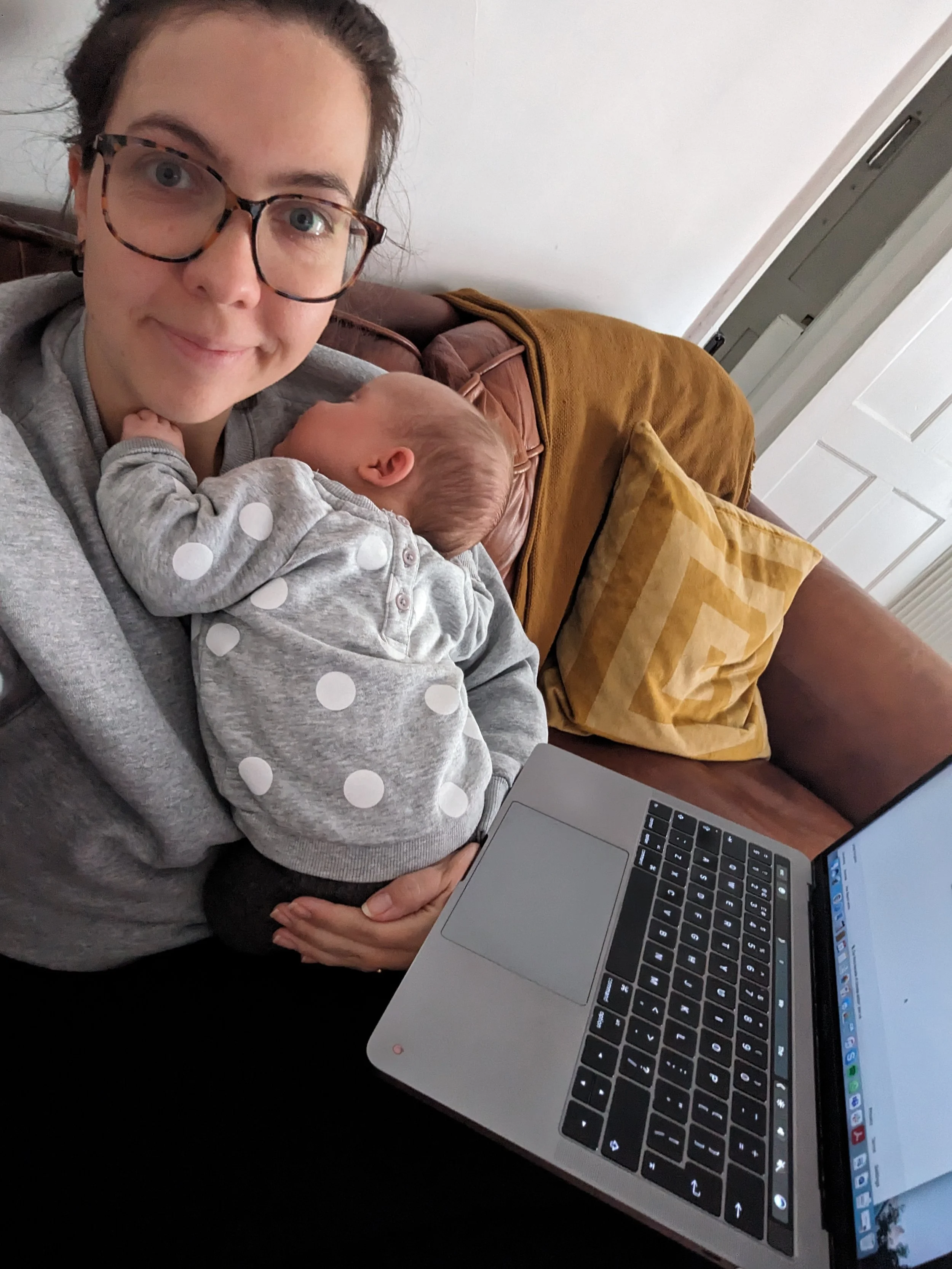 A woman with glasses and a gray sweater holding a sleeping baby with a polka-dot outfit, sitting on a couch with yellow pillows and a brown blanket, working on a silver laptop in a room with a white wall and a door.