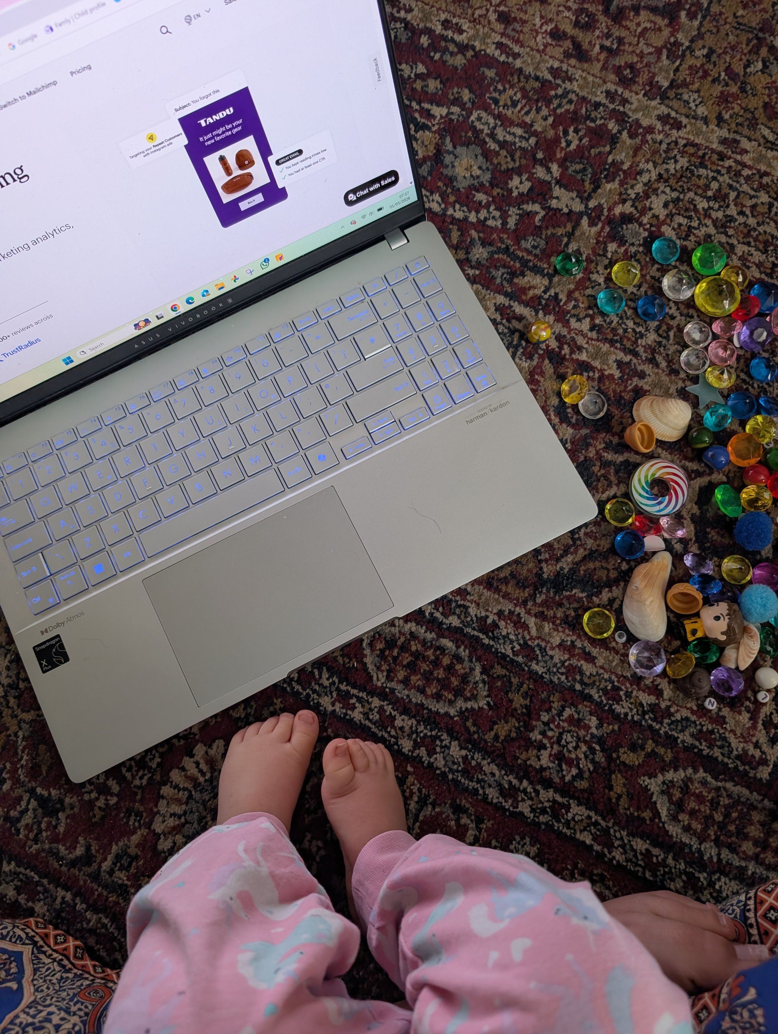 Child in pink pajamas holding adult's hands with a silver laptop on a patterned rug nearby. Colorful glass marbles are scattered on the rug.