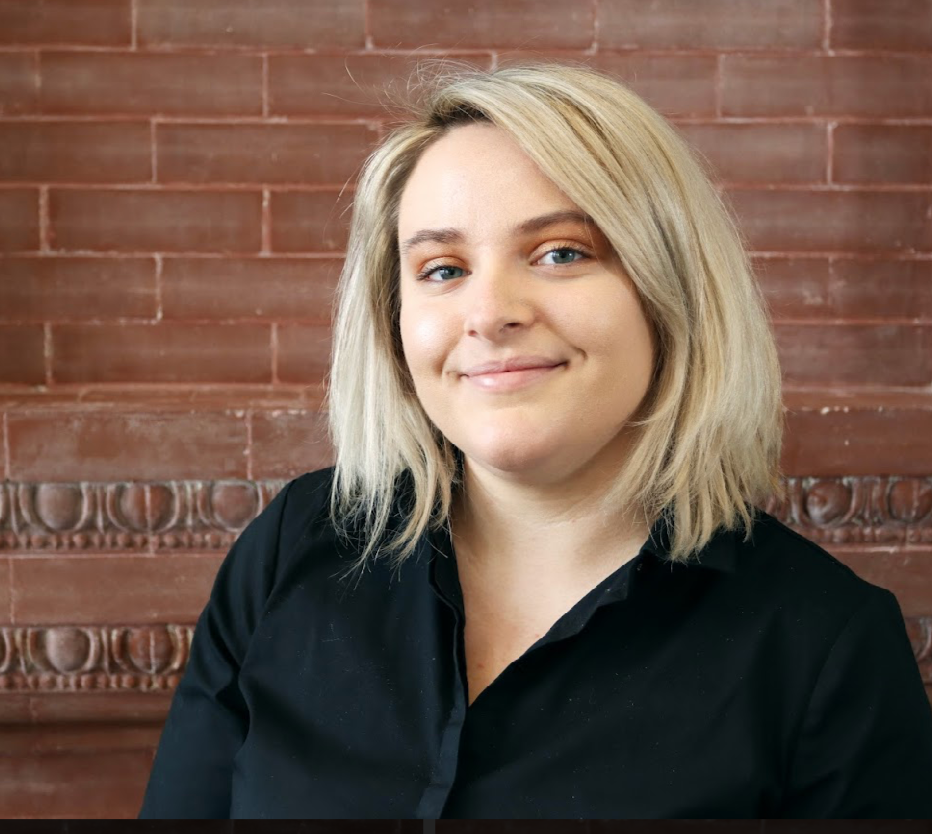 Head-and-shoulders portrait of a young woman with shoulder-length blonde hair and blue eyes, smiling slightly, against a red brick wall background.
