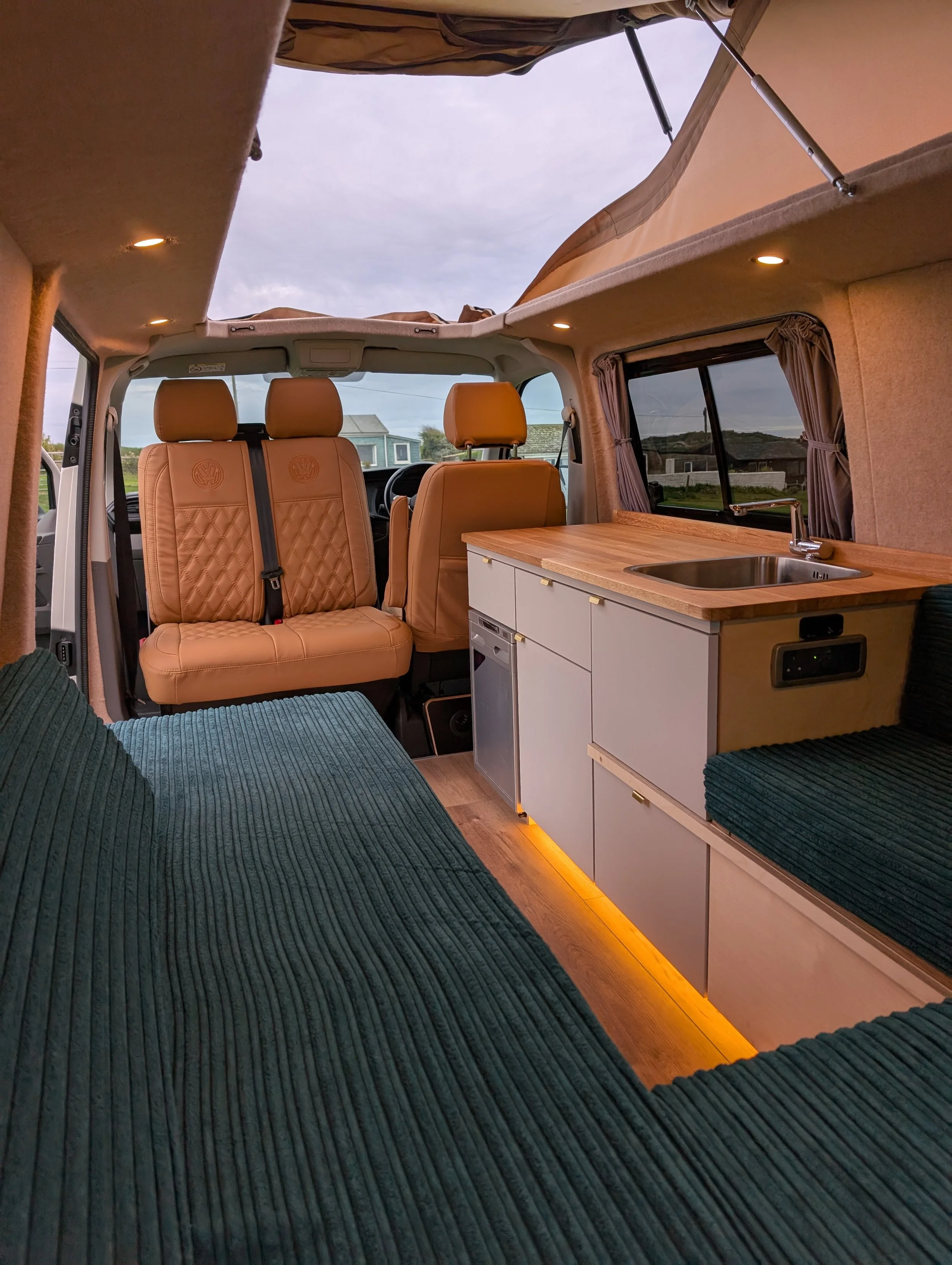 Interior view of a camper van with tan leather seats, a small kitchenette with a wooden countertop and sink, and windows with curtains. The ceiling has small recessed lights, and the sky is visible through an open roof hatch.