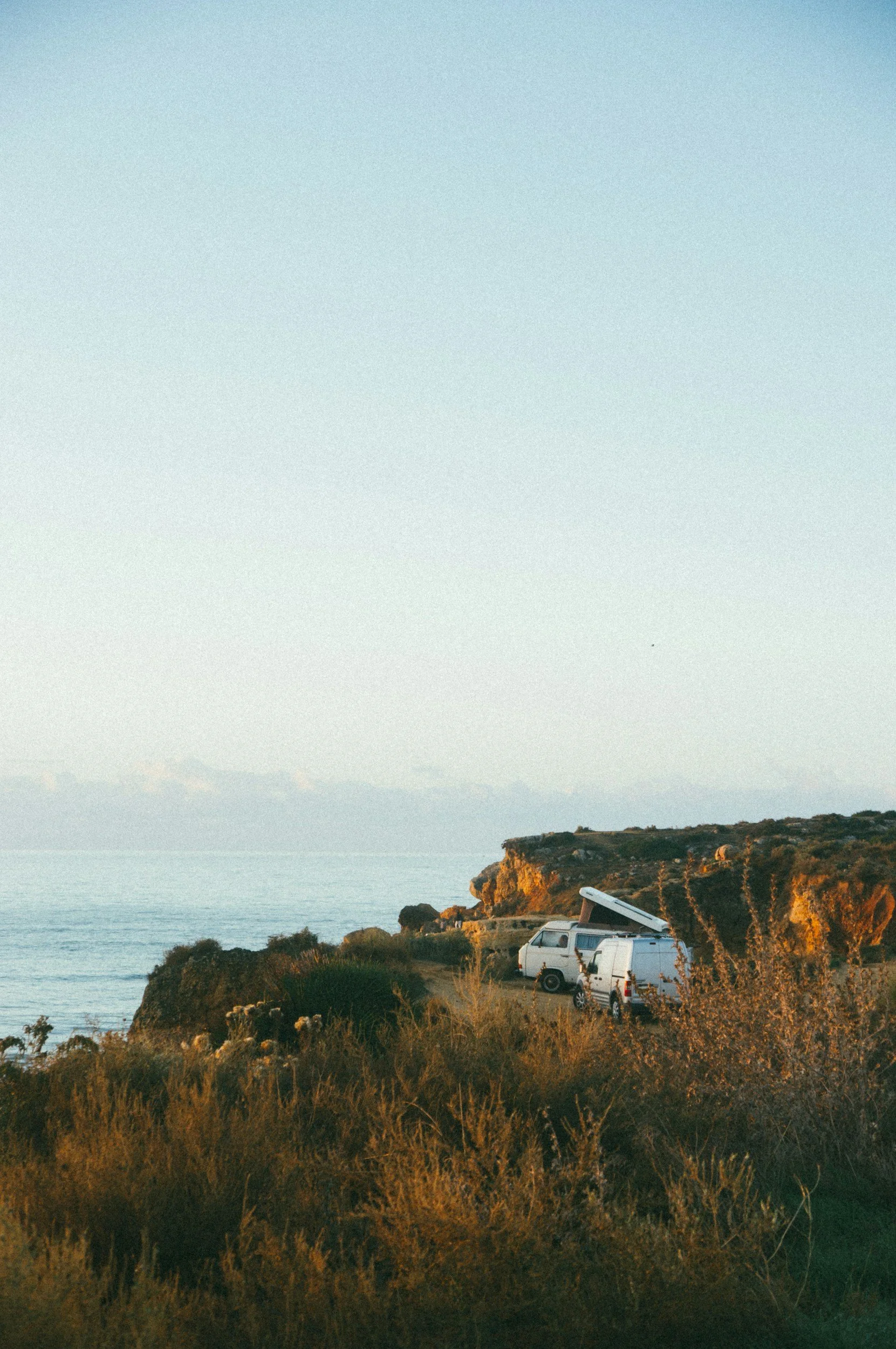 Beachside landscape with a cliff, ocean, and parked vans, one of which has an awning extended, during sunset.