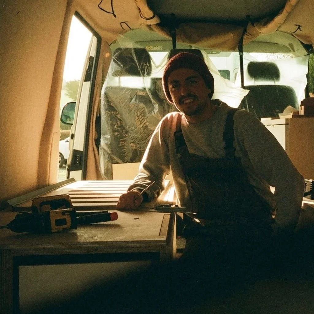 A young man with a beard and mustache, wearing a red beanie, smiling and sitting inside a van with a table and tools around him.