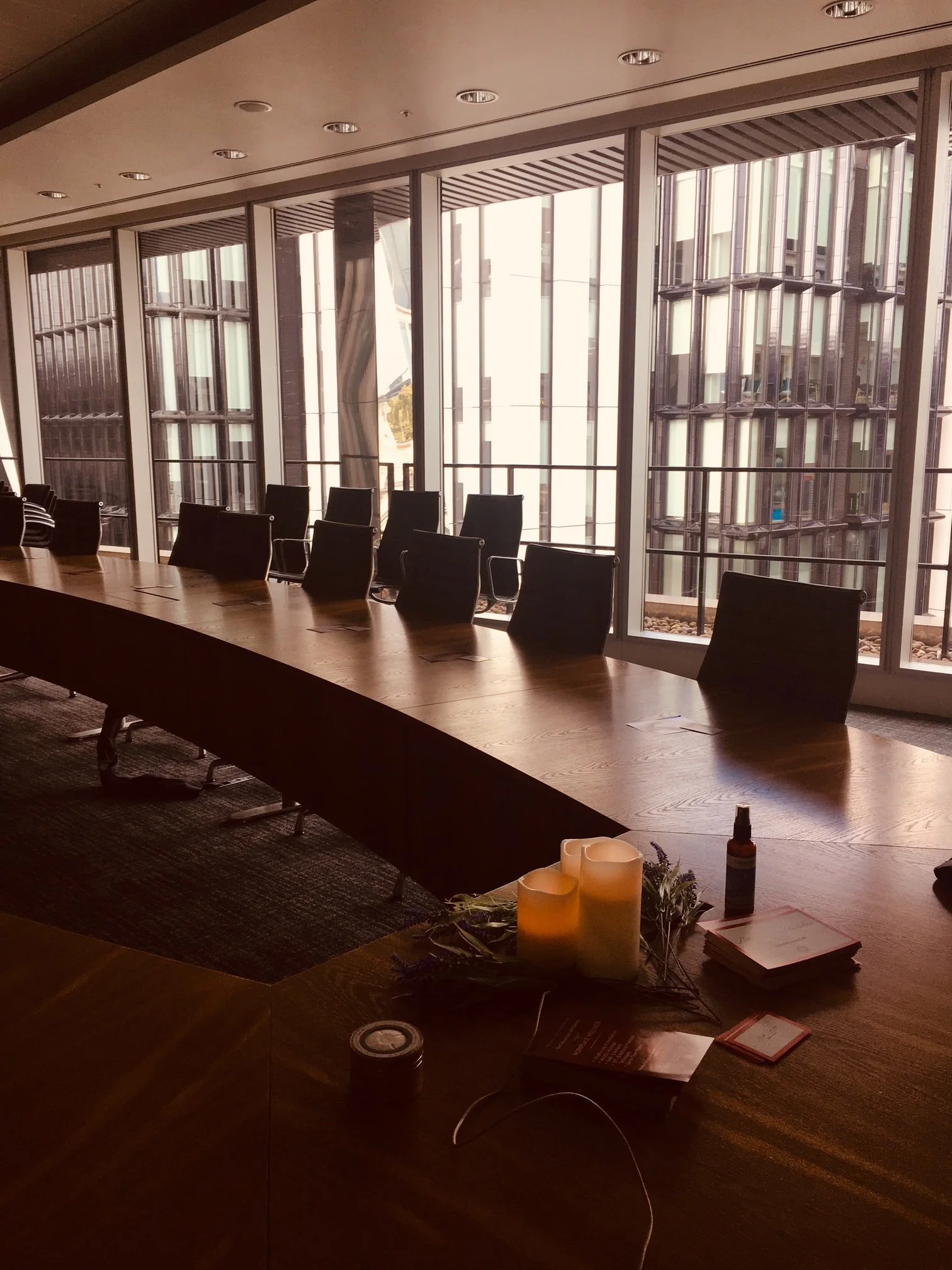 A conference room with a long wooden table and black chairs, large windows with views of tall buildings outside, candles and decorative items on the table, and candles and papers in the foreground.