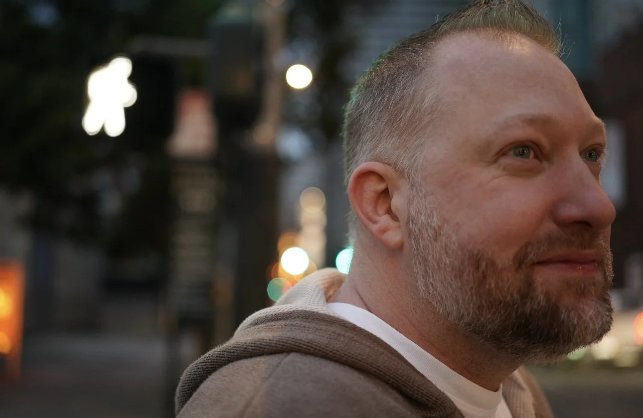 Close-up of a smiling man with facial hair, short hair, wearing a hoodie, on a city street at dusk, with blurred street lights and pedestrian signals in the background.