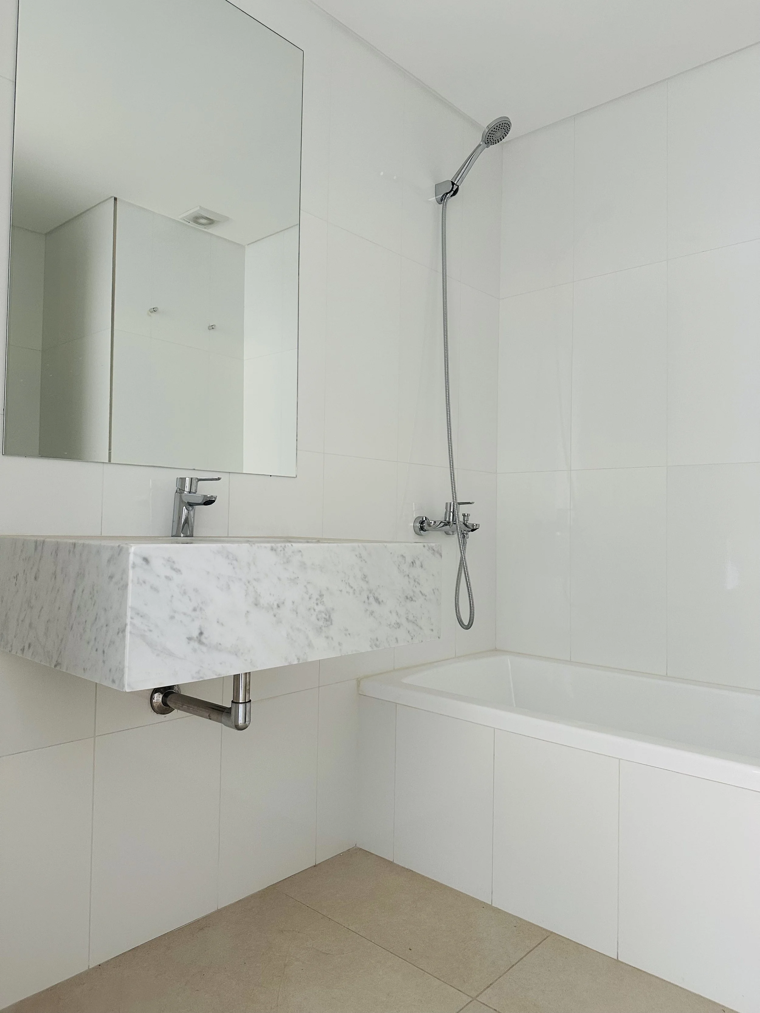 Venice Tigre. Temporaty apt. A minimalist bathroom with white tile walls, a white bathtub, a marble countertop sink, a wall-mounted mirror, and a handheld showerhead.