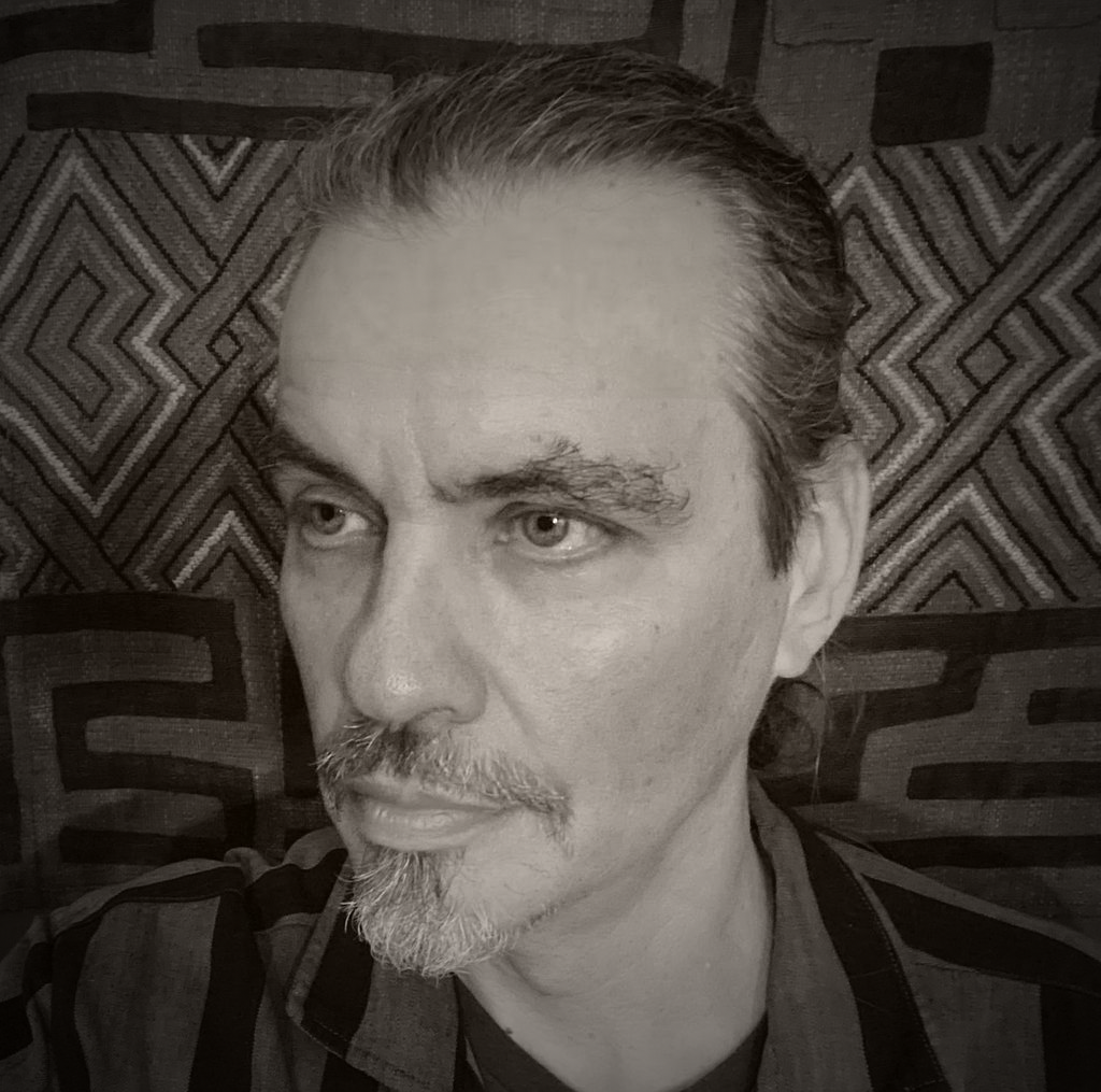 A black and white portrait of a man with slicked-back hair and a goatee, sitting in front of a patterned fabric background.