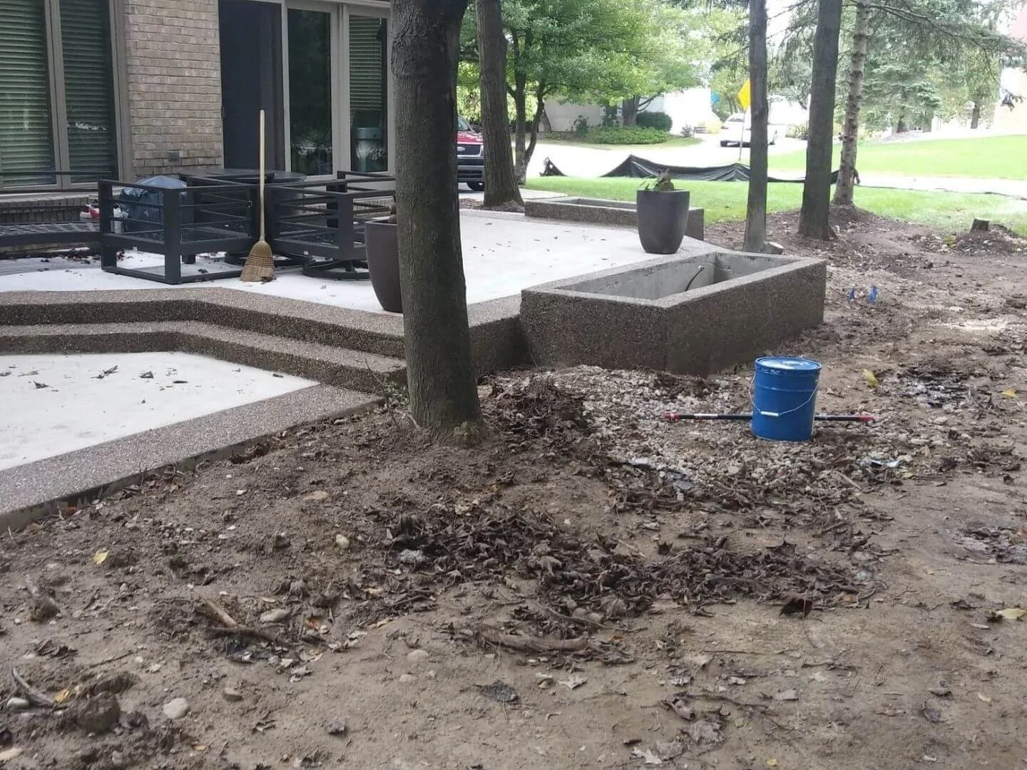Construction site in a backyard with steps leading to a patio, a empty planters, a bucket, and a broom leaning against the railing. Dirt and leaves in the foreground with trees and grass in the background.
