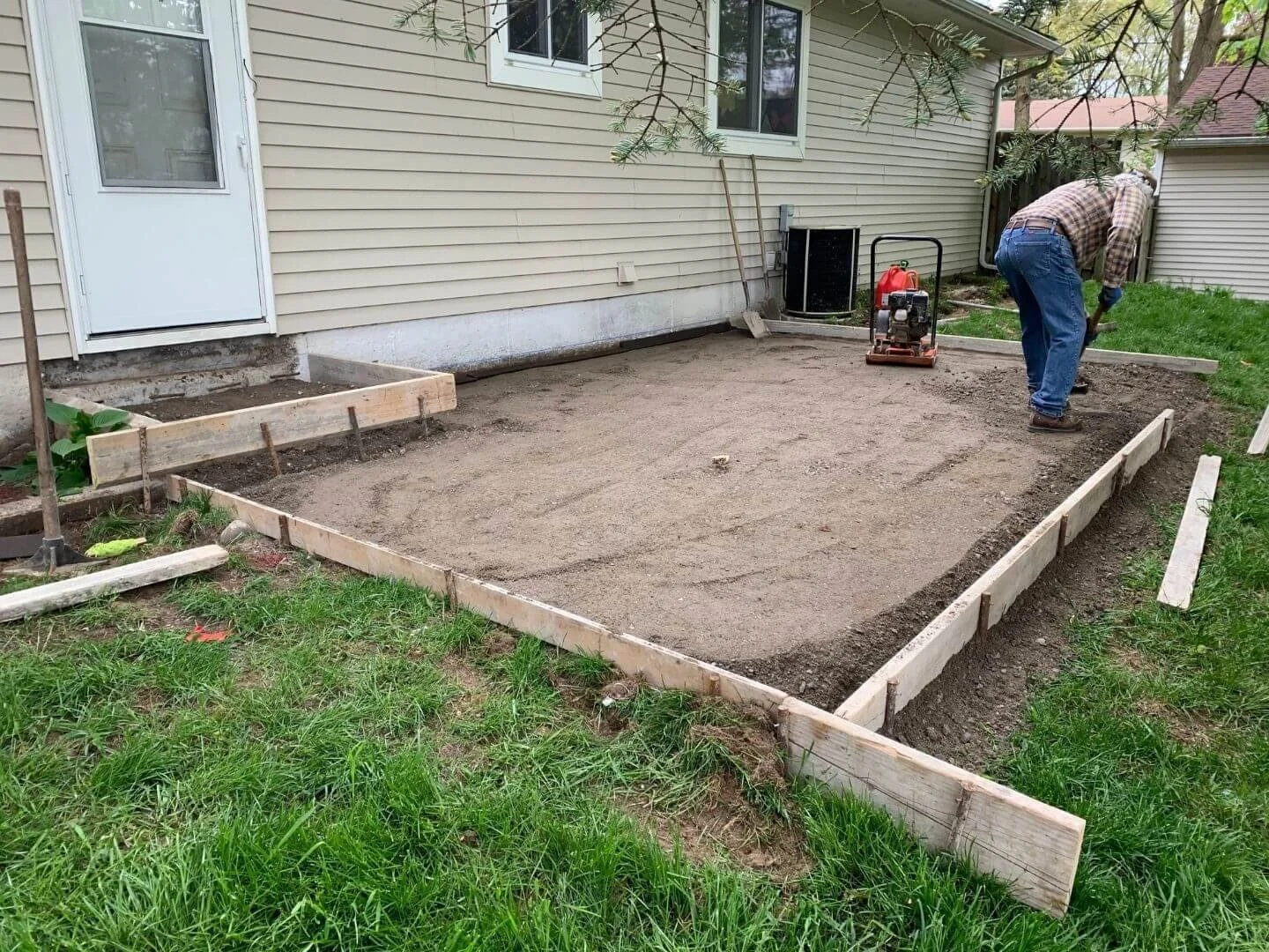 A person working on a backyard patio project, leveling and preparing the ground with wooden framing around a dirt area near a house.