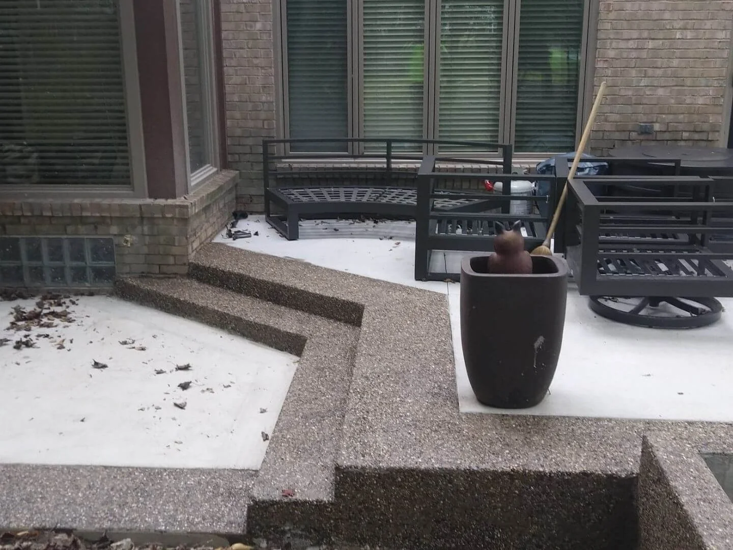 Backyard patio with black metal benches, a broomstick in a large pot with a cat face, and a brick wall with large windows.