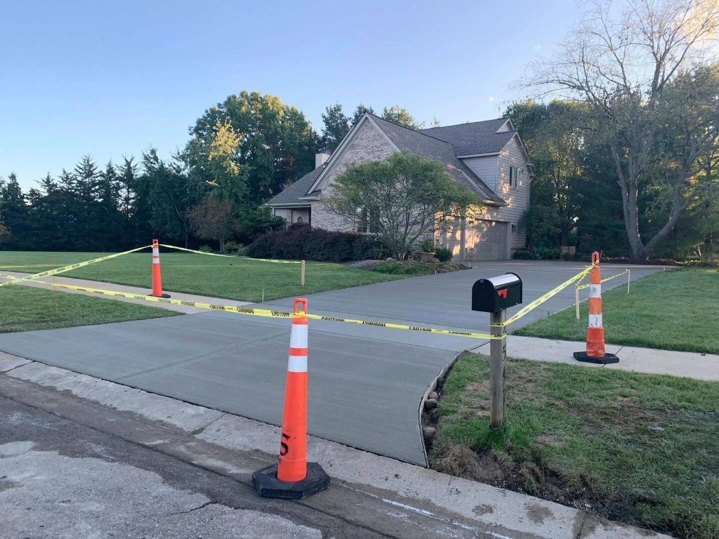 Residential driveway with new concrete fresh pour, surrounded by orange construction cones and caution tape for drying and setting, with a mailbox in the foreground and a house with trees in the background.
