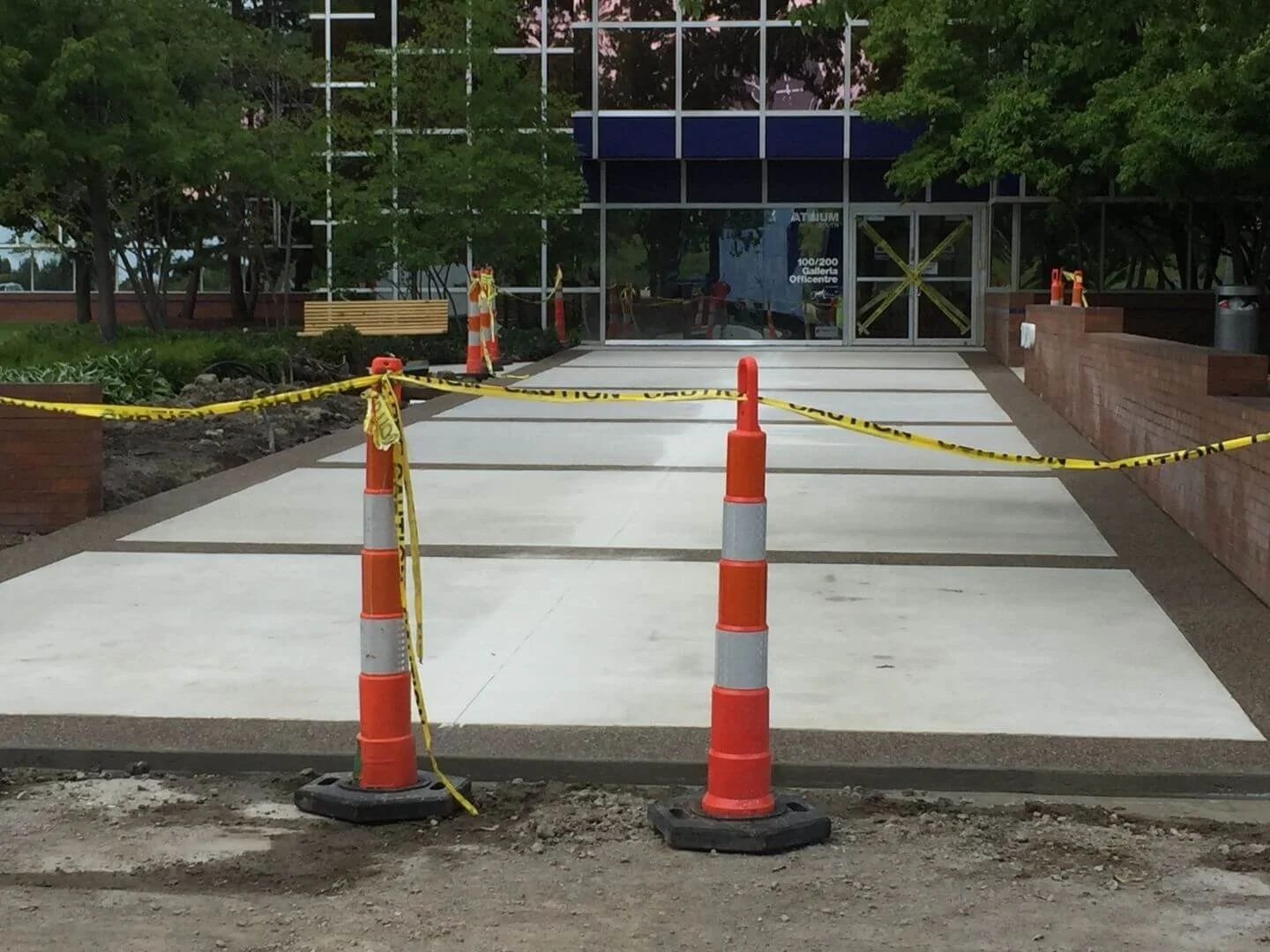 Construction area with orange traffic cones and yellow caution tape, sectioning off a newly poured concrete sidewalk in front of a glass building entrance, surrounded by trees.