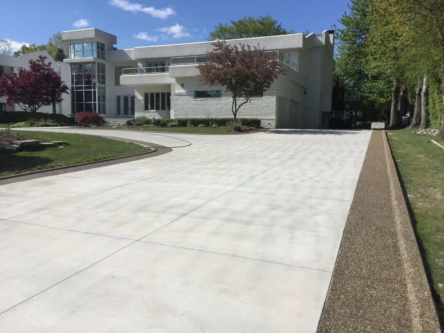 Modern white multi-story house with large glass windows and a driveway, surrounded by green trees and landscaped lawn on a sunny day.