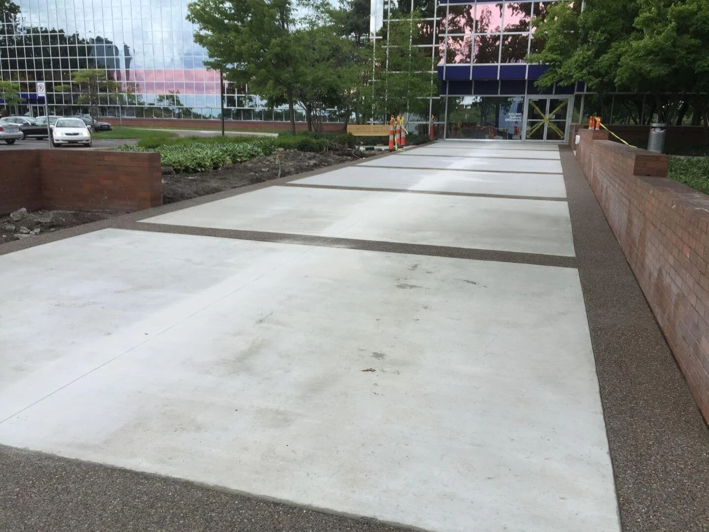 Newly constructed concrete parking lot with designated parking spaces separated by dark bands, flanked by brick planters and green trees, outside a glass building.