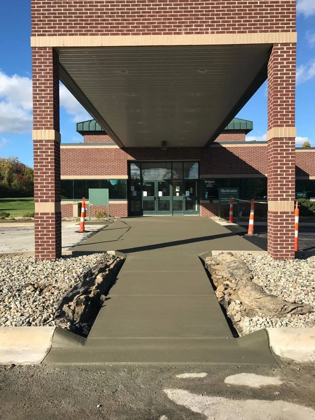 New sidewalk leading to the entrance of a brick building with glass doors. Construction cones are placed on either side of the sidewalk, which is partially unfinished. The building has a sign with the name 'McLaren'.