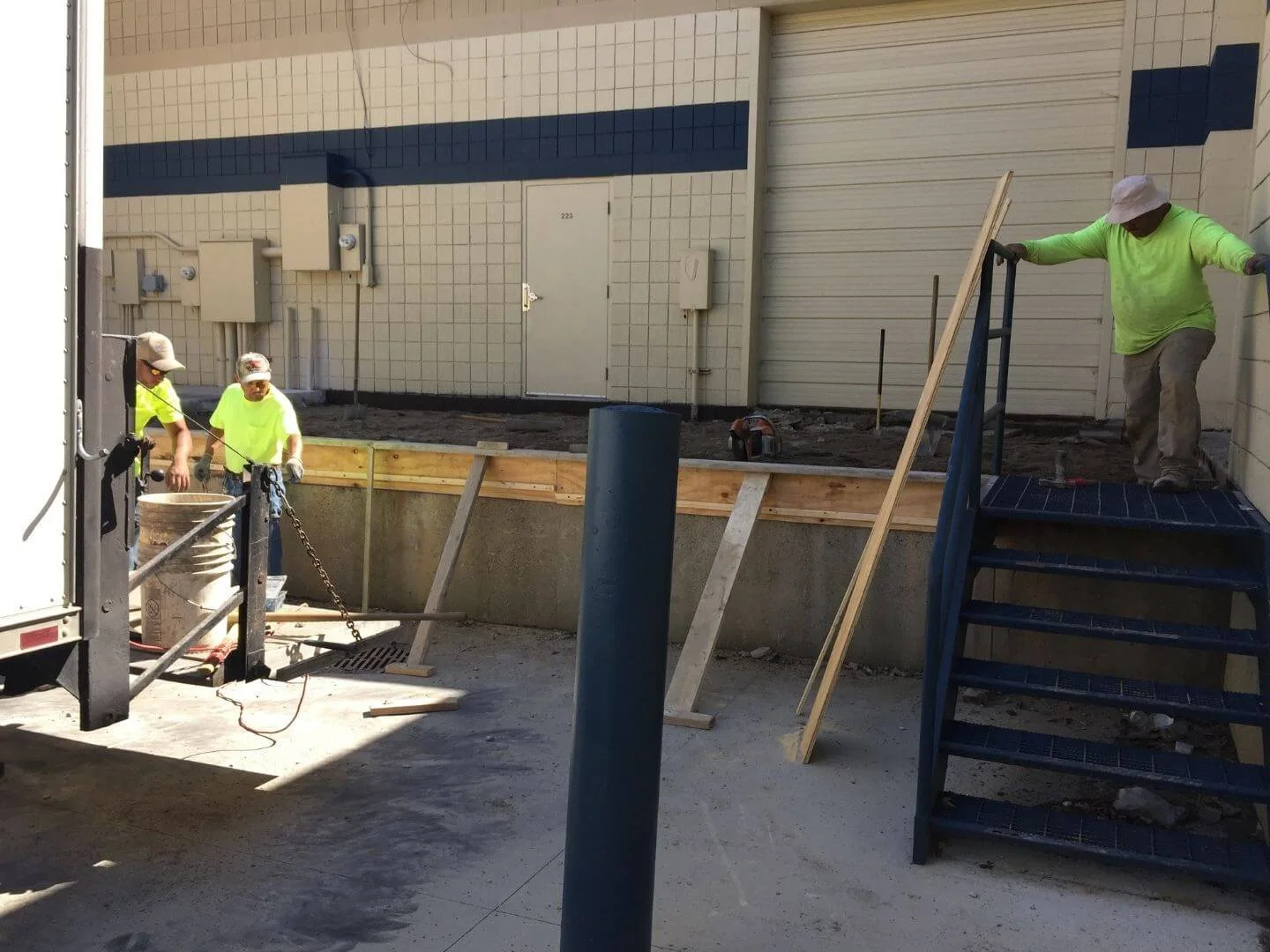 Construction workers wearing yellow shirts and hats working on an outdoor building project, with some standing on the ground and one on stairs, surrounded by construction materials and tools.