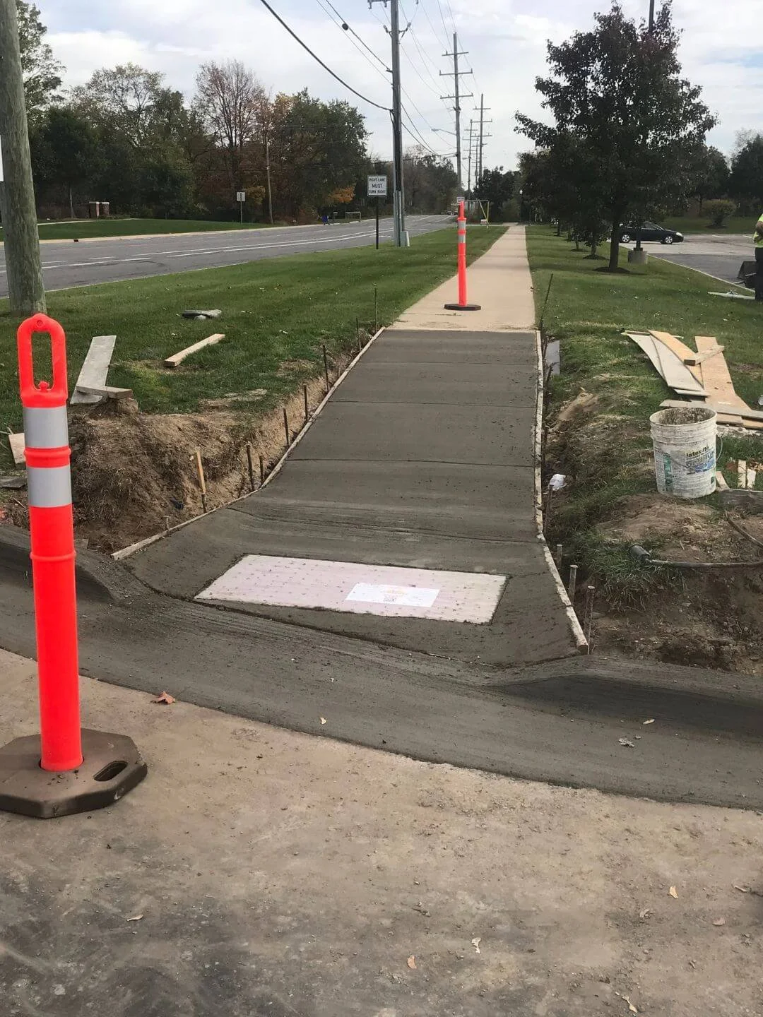 Newly constructed sidewalk and curb under construction with orange safety cones and barriers, adjacent to a grassy area and road with power lines.