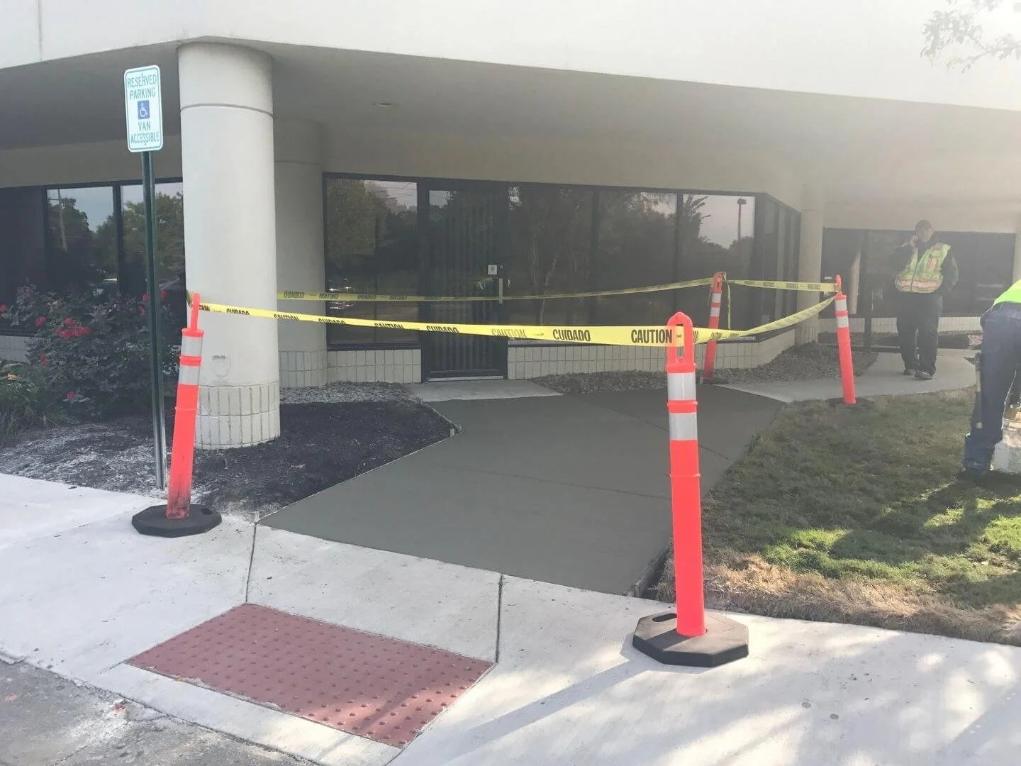 Sidewalk leading to a building entrance with new concrete, surrounded by orange cones and caution tape, and two workers wearing safety vests nearby.