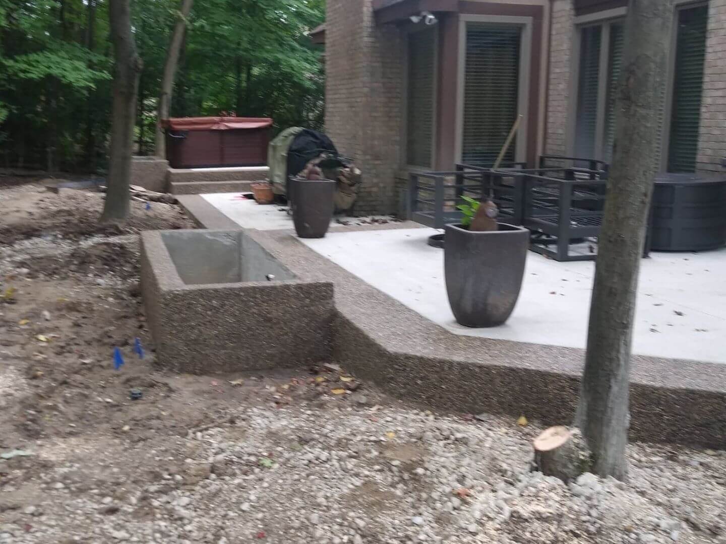 Newly constructed backyard patio with concrete flooring, potted plants, a hot tub, and trees surrounding the area.
