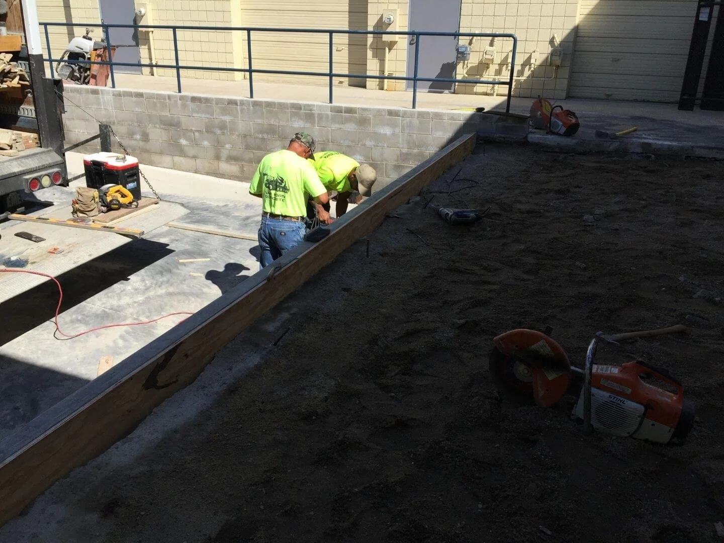 Two construction workers in neon yellow shirts working on a sidewalk or driveway, with various tools and construction equipment around them, and a rising wall in the background.
