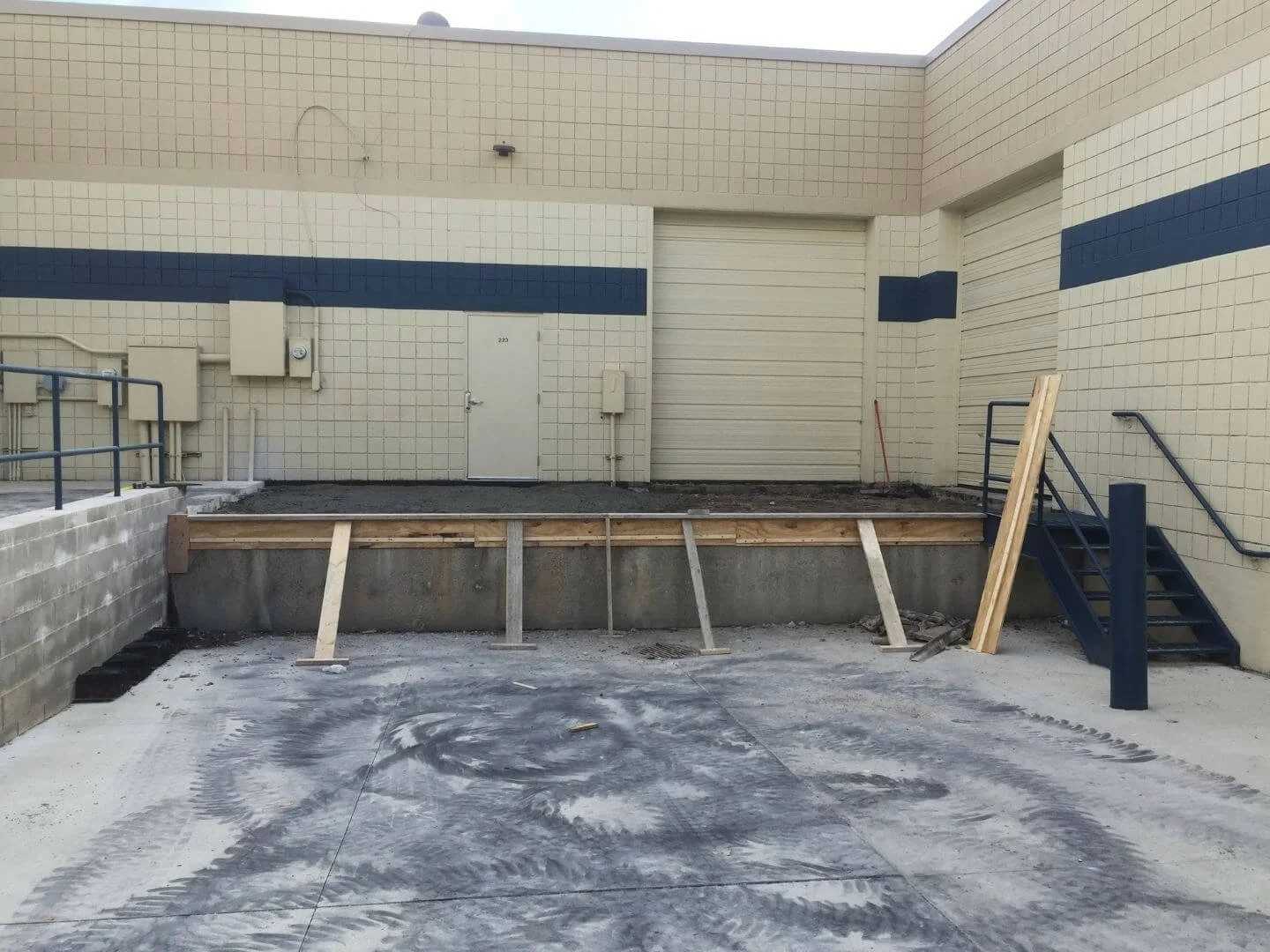 Construction site with a raised platform, wooden support beams, stairs on the right, and a beige building wall with a door and closed roll-up door.
