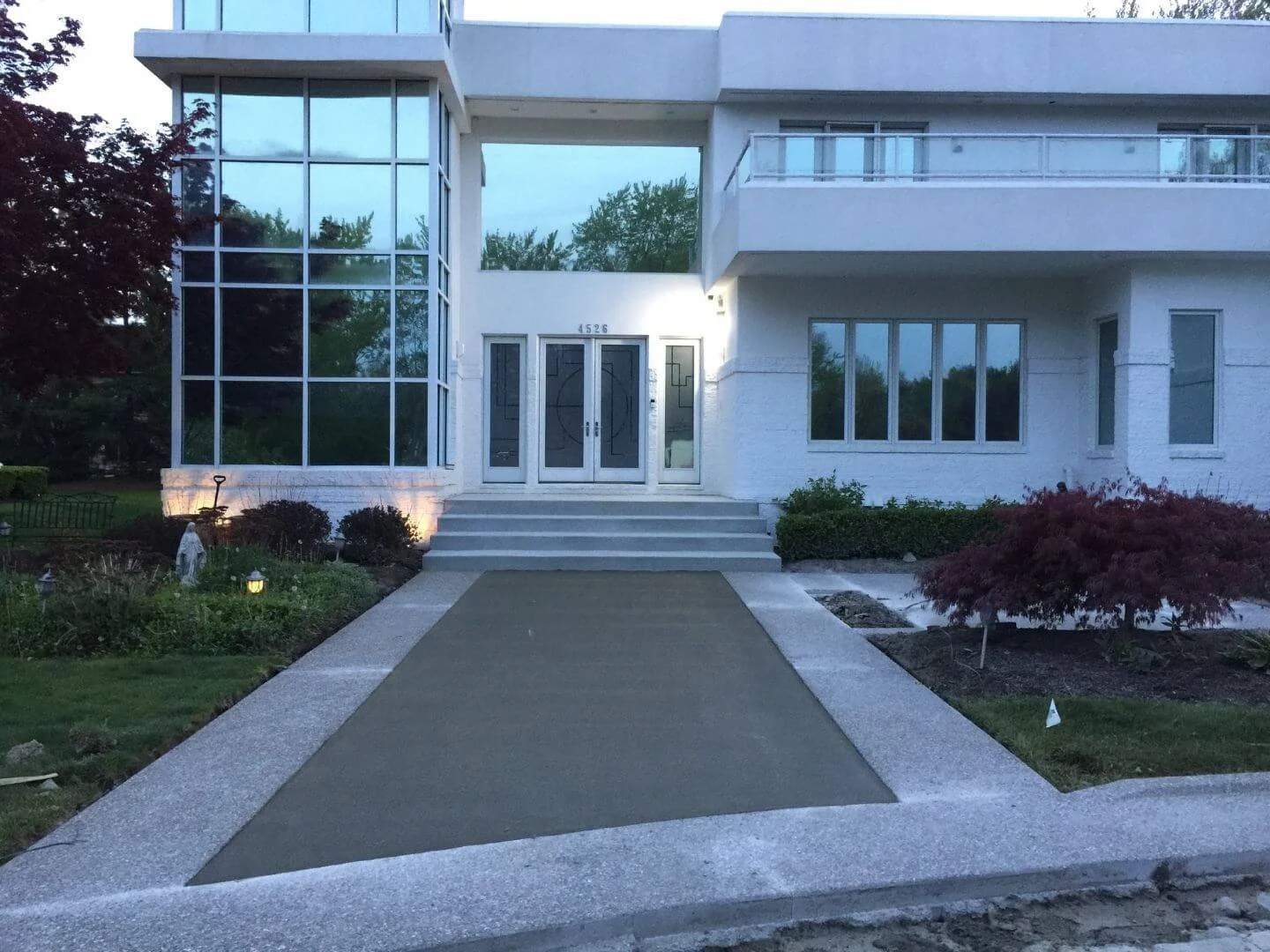 Front entrance of a modern white building with glass windows, steps leading up to black double doors, and a landscaped garden with bushes and trees.