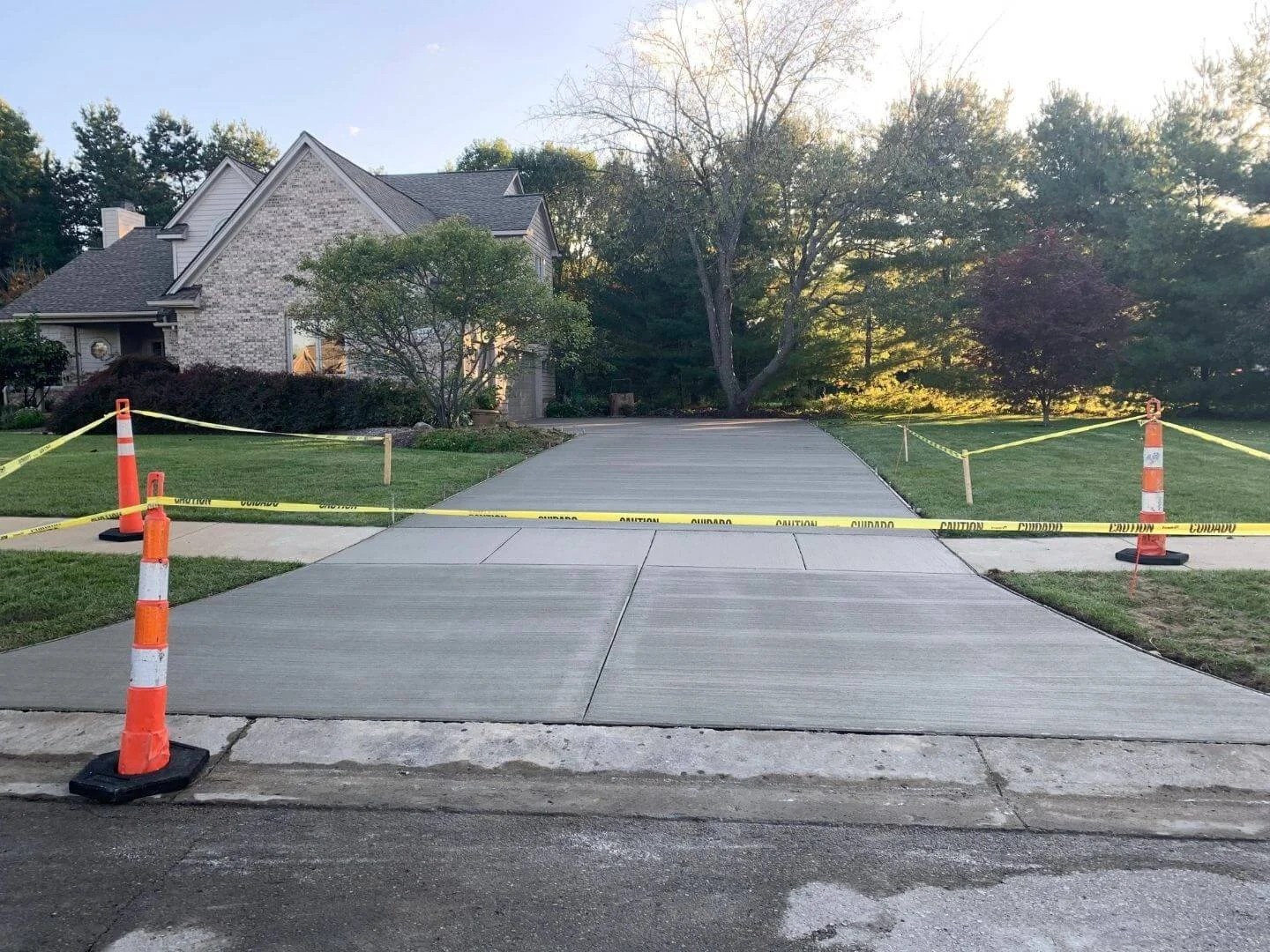 Newly paved driveway blocked off with yellow caution tape and orange traffic cones in front of a house with trees and a lawn.