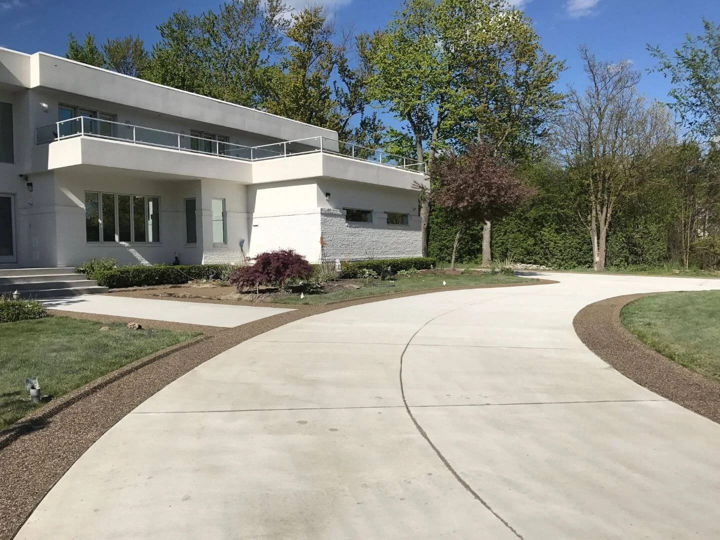 Modern white house with large windows and a curved driveway, surrounded by trees and landscaped yard with shrubs and grass.