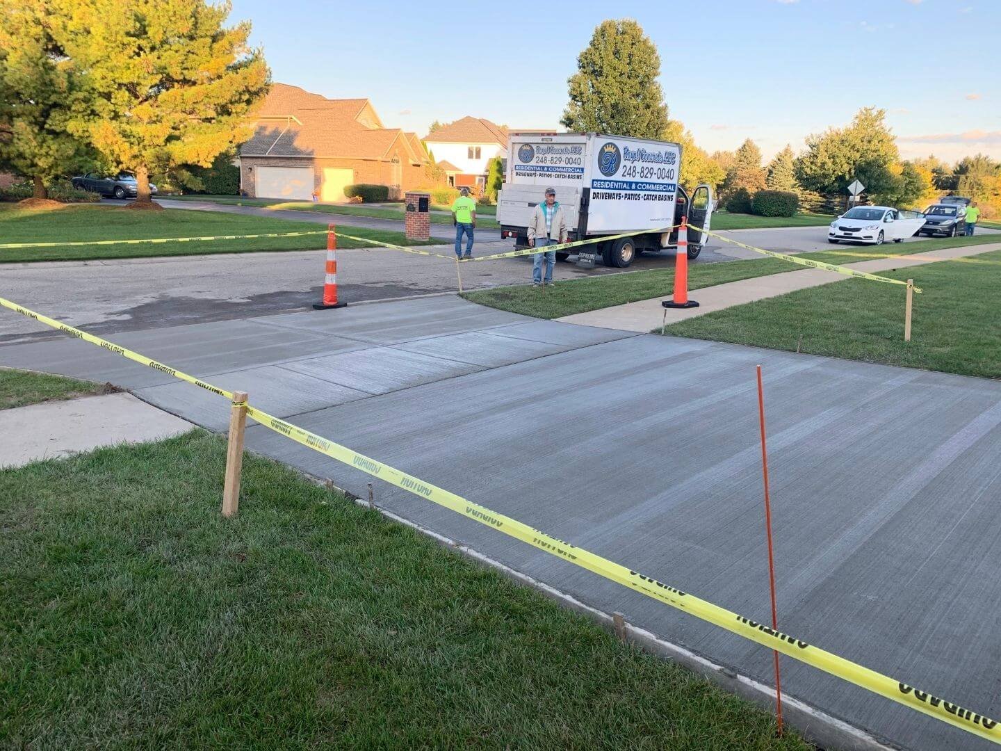 Workers installing a concrete driveway in a residential neighborhood, surrounded by yellow caution tape and orange traffic cones, with houses, trees, and a clear sky in the background.